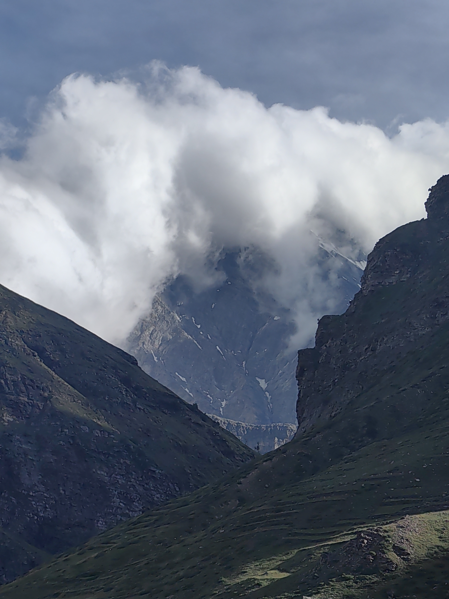 The image depicts a majestic mountain landscape, likely in the vicinity of Yamling, India. The foreground is dominated by steep, verdant slopes covered in grass and scattered rocks. These slopes converge to form a valley. In the midground, a rugged, rocky peak is partially obscured by dramatic, billowing white clouds that also frame the sky. Beyond this, a snow-capped mountain rises, its rugged texture visible through breaks in the clouds. The sky is a pale blue, with hints of grey in the clouds. The overall impression is one of a remote, wild, and breathtaking natural environment.