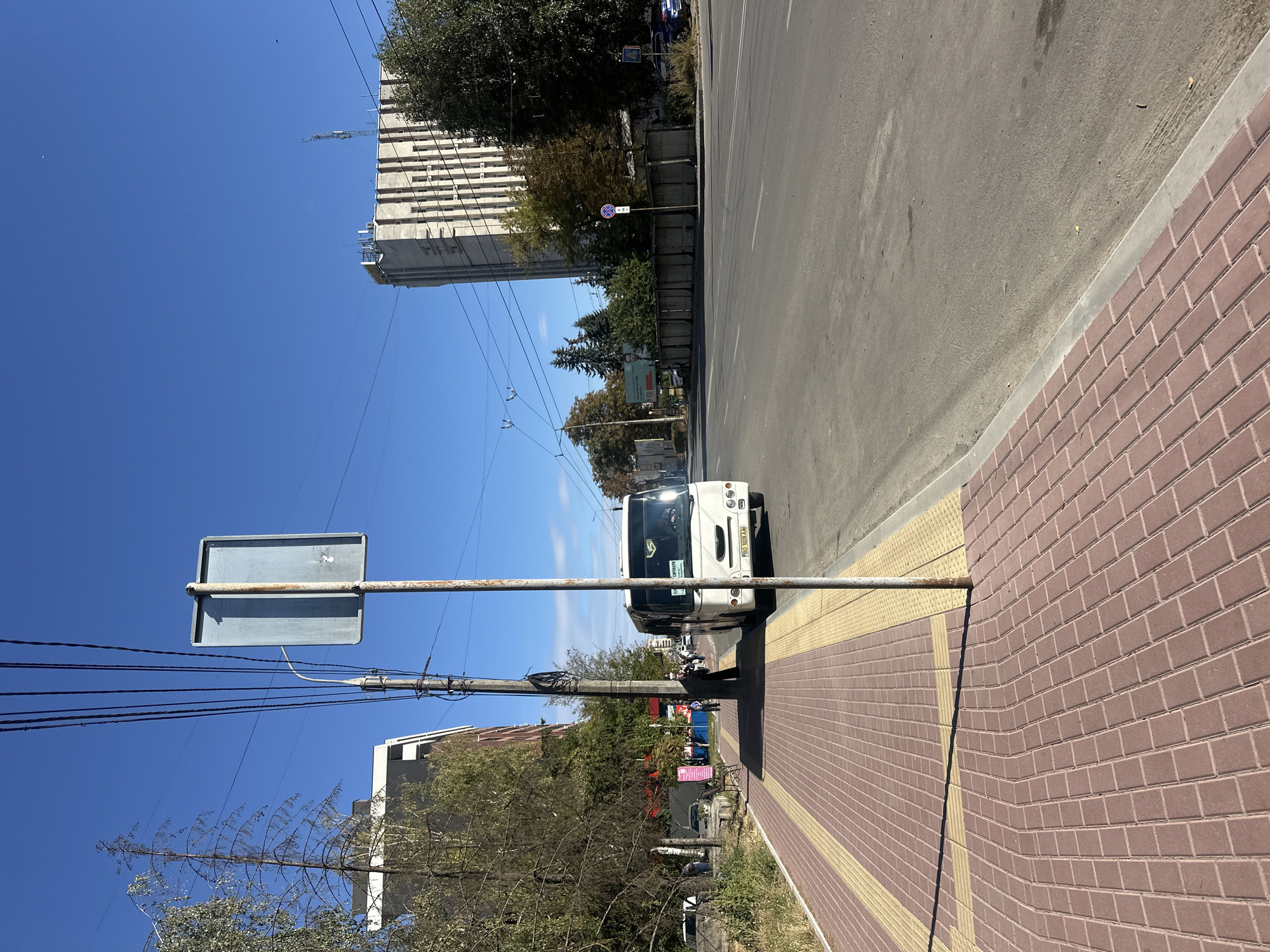 The image captures a street scene in Ternopil, Ukraine, under a clear blue sky. On the right side of the frame, a white bus is parked on the asphalt road. In the foreground, a red brick sidewalk runs along the left side of the road, with a yellow tactile paving strip bordering the curb. The sidewalk appears to be made of interlocking bricks, some of which are slightly warped. A metal pole with a weathered sign stands on the sidewalk.
To the left of the sidewalk, there are trees and a multi-story building. Further down the street, another tall building with a distinctive facade and antennas on its roof is visible. In the middle ground, there are more trees and a smaller building. Several people are walking on the sidewalk. The overall scene suggests a typical urban environment on a bright, sunny day.