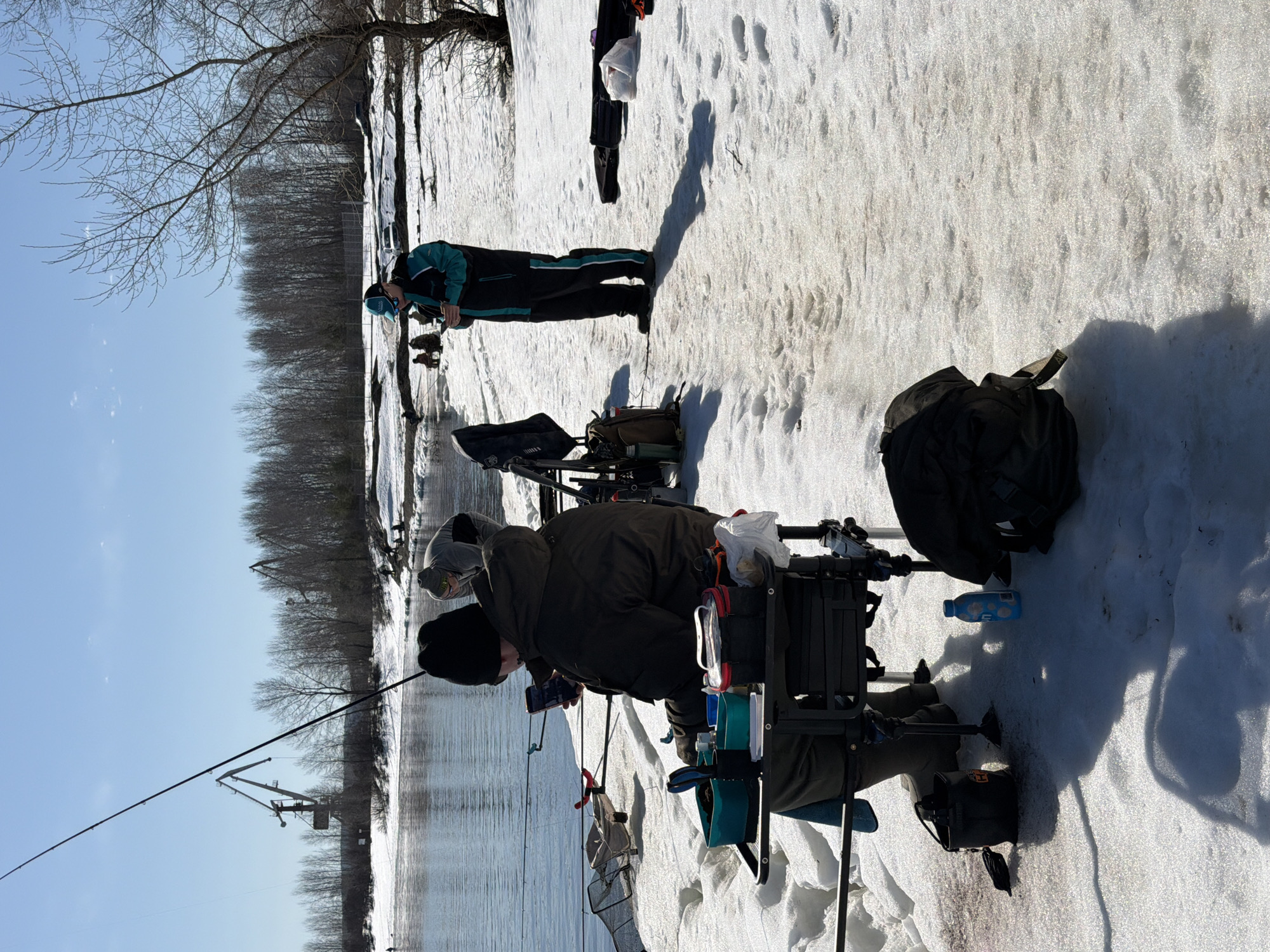 Two people are fishing on a snowy bank beside a river. The person closest to the camera is seated, with fishing equipment surrounding them. The person further away is standing. Cranes are visible across the river. The location is Noviy, Russia.