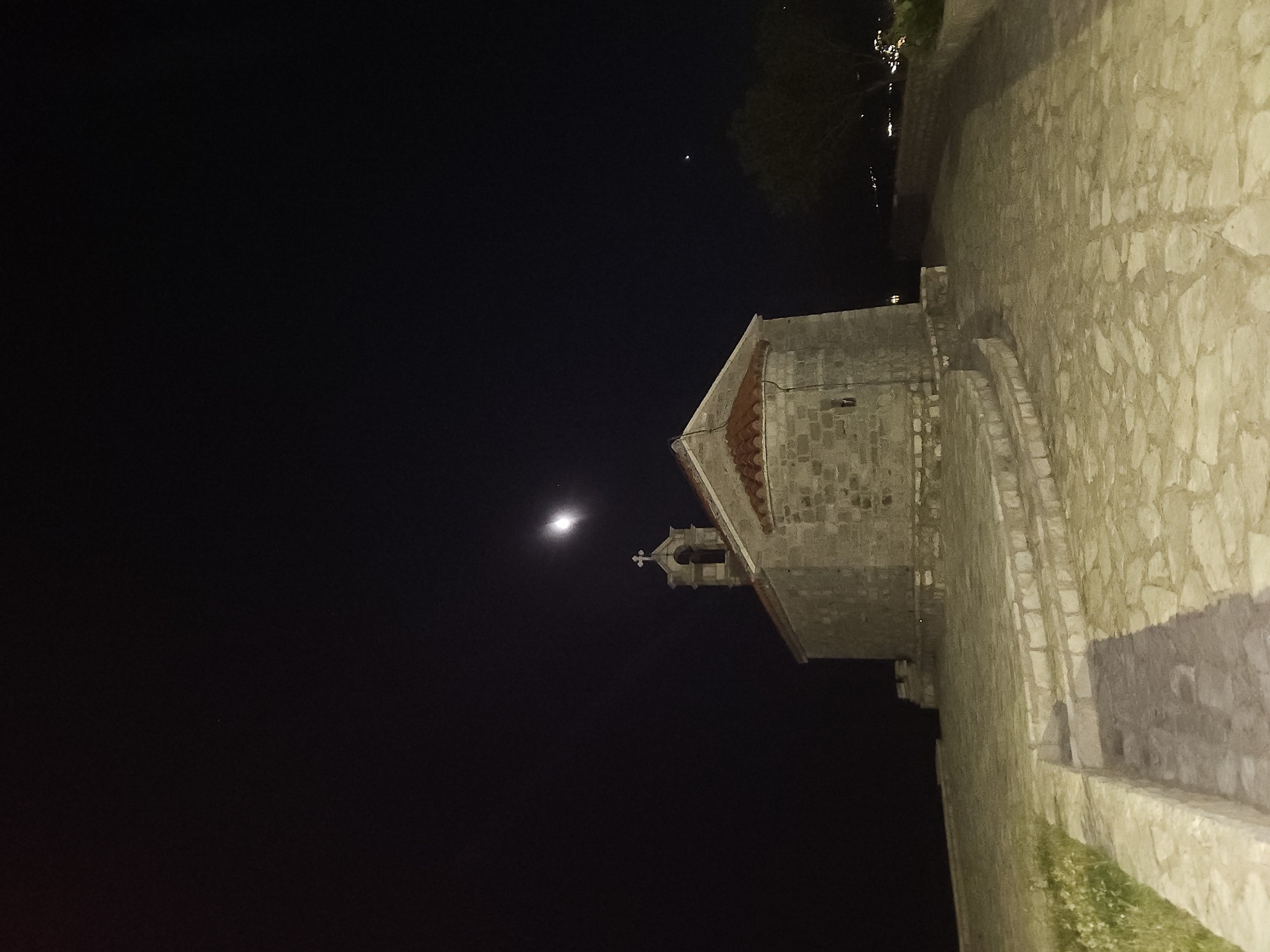 The image shows a small, stone church with a red-tiled roof at night. The church has a bell tower with a cross on top. The church is situated on a stone-paved area, with steps leading up to it. There is a faint moon or a bright star visible in the dark night sky. To the right of the church, there is a silhouette of a tree and some distant lights, suggesting a settlement. The scene appears to be on an elevated area, possibly a hill or a promontory, overlooking the sea. This location is the Church of Saint Sava on the Sveti Stefan islet in Montenegro.