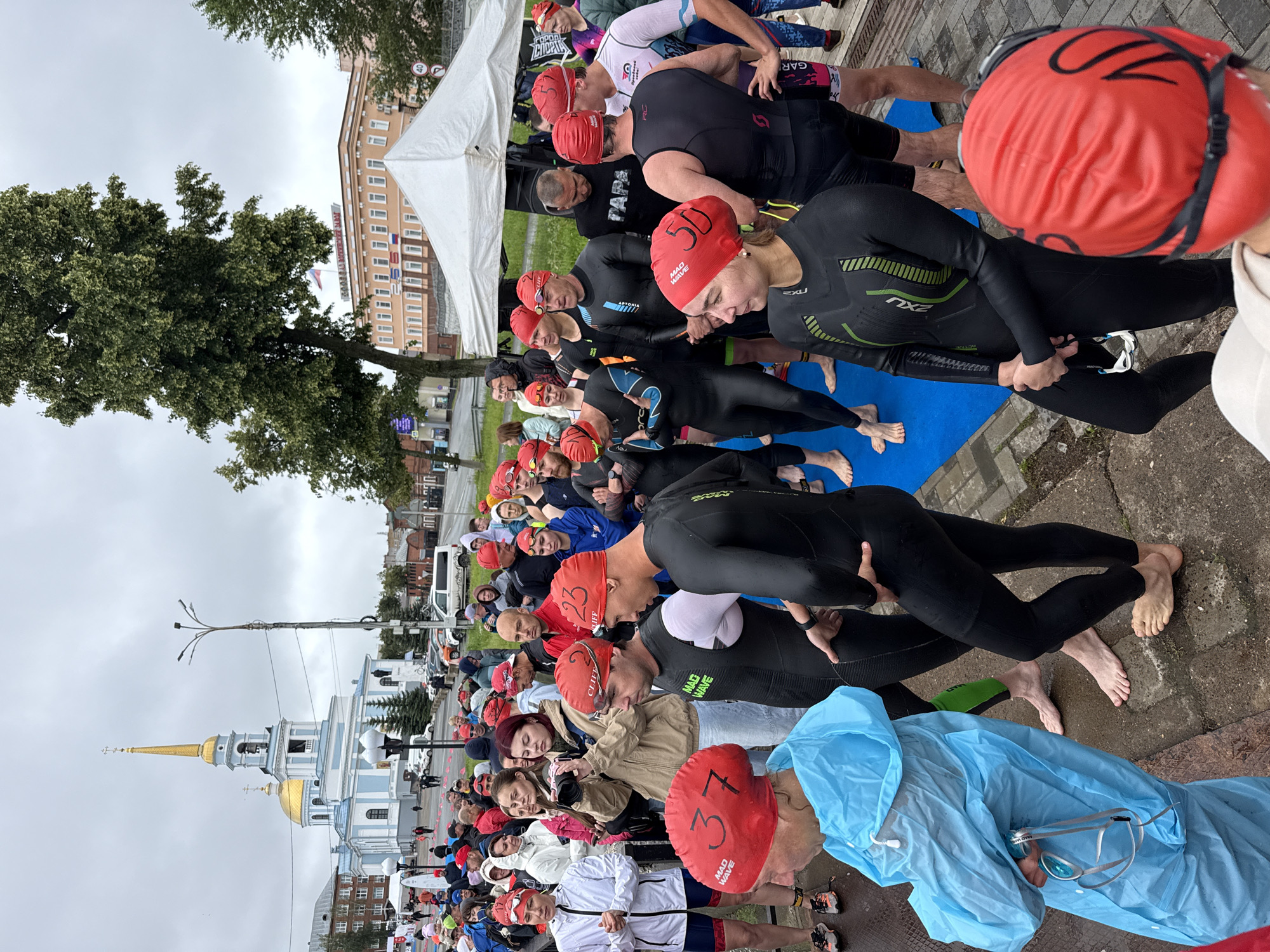 The image depicts a gathering of people, likely participants in an outdoor sports event, in the city of Votkinsk, Russia. In the background, a prominent light blue church with a golden dome and spire stands against an overcast sky. Several individuals, predominantly wearing red swim caps, are positioned in the foreground and midground. Some are wearing wetsuits, suggesting a swimming or triathlon event. One person in the foreground is wearing a bright blue raincoat and holding a pair of swimming goggles. The ground is paved with concrete and a blue mat is laid out, possibly for the event. Buildings, trees, and streetlights are visible in the surrounding environment. The overall atmosphere appears to be one of preparation or anticipation for an event.