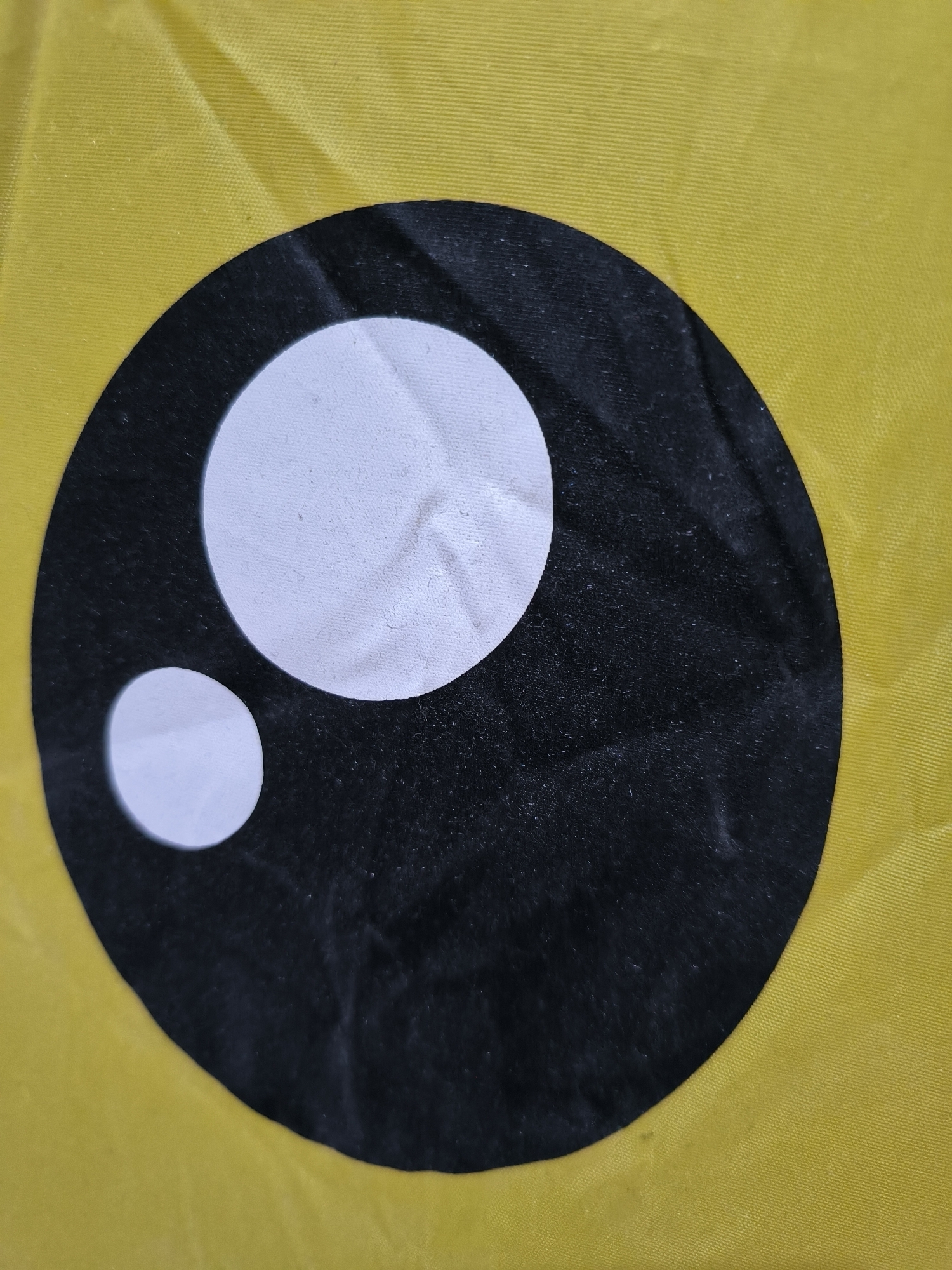 The image shows a close-up of a yellow fabric with a cartoon eye design on it. The eye consists of a large black circle with two white circles inside representing the pupil and a secondary highlight. The fabric appears slightly wrinkled and textured. The location is not discernible from the provided image.