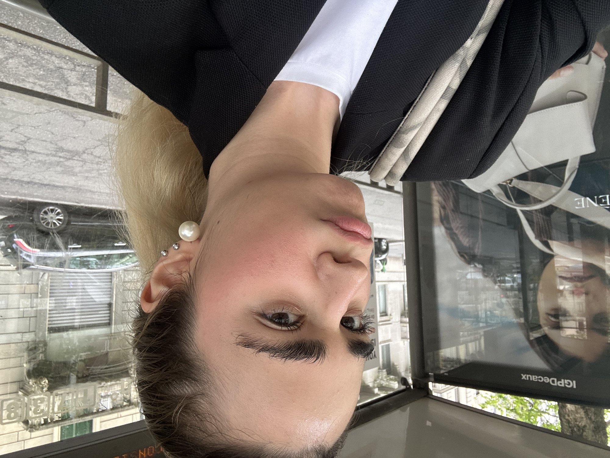 A vertically-oriented medium shot depicts a woman standing near an advertisement display in an urban environment. The advertisement display features the text "IGPDecaux". The woman has fair skin, dark eyebrows, and light blonde hair pulled back. She wears a pearl earring. She is dressed in a black blazer over a white top. Reflected in the glass of the advertisement display are buildings, a car, and trees, suggesting a street scene. The overall setting suggests a metropolitan area. Due to the "IGPDecaux" advertisement, the location is the city of Milan, Italy.