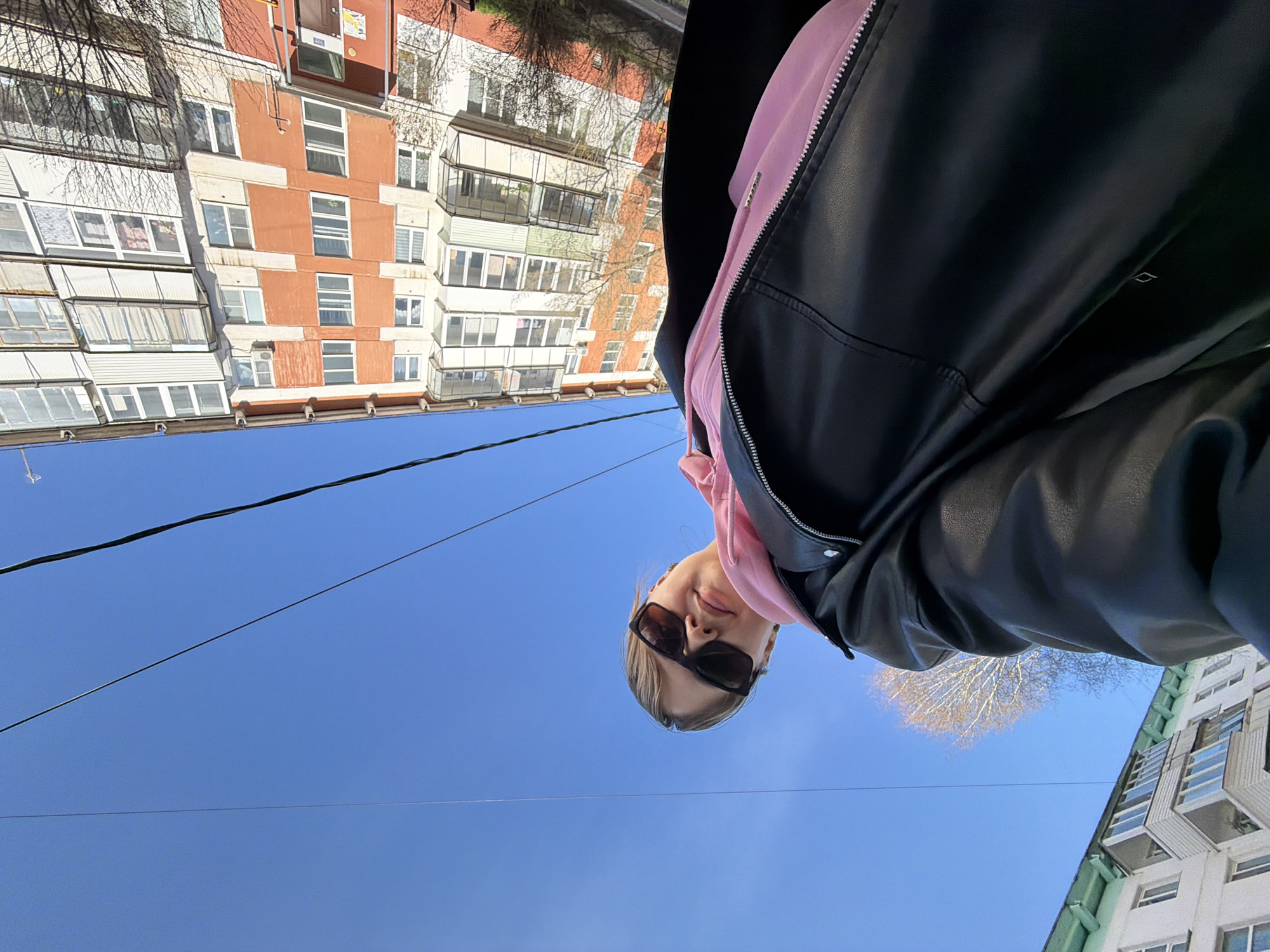 A low-angle outdoor portrait features a young woman looking down towards the camera, positioned against a clear blue sky and urban backdrop. The woman, likely in her late teens or early twenties, has light-colored hair and wears dark, rectangular sunglasses. Her expression appears neutral, with a slight hint of a smile. She is dressed in a pink hooded sweatshirt beneath a black leather jacket, with a silver zipper visible on the jacket.
In the background, two multi-story residential buildings frame the scene. The building on the left features a facade with alternating sections of reddish-orange and light cream or white, punctuated by numerous windows and enclosed balconies. The building on the right is partially visible, appearing lighter in color with some greenish accents. Bare tree branches, suggesting a late winter or early spring season, are visible near both buildings.
Several dark power lines stretch horizontally across the bright, cloudless blue sky, dividing the upper and lower portions of the background. The lighting indicates a clear, sunny daytime. No legible text is visible within the image.