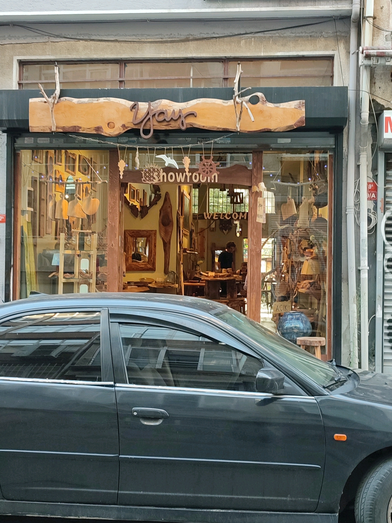 A dark gray car is parked in front of a storefront in Kadıköy, Turkey. The storefront appears to be a workshop or showroom, with various wooden items displayed inside and hanging from the ceiling. A sign above the entrance reads "YAY showroom". The car is a sedan, and its windshield is visible in the foreground. The storefront has large glass windows that offer a view of the interior. There are a few people visible inside the shop, appearing to be working or browsing. The street outside is paved, and there is a building across the street with a red and white sign. The overall scene depicts a typical urban streetscape.