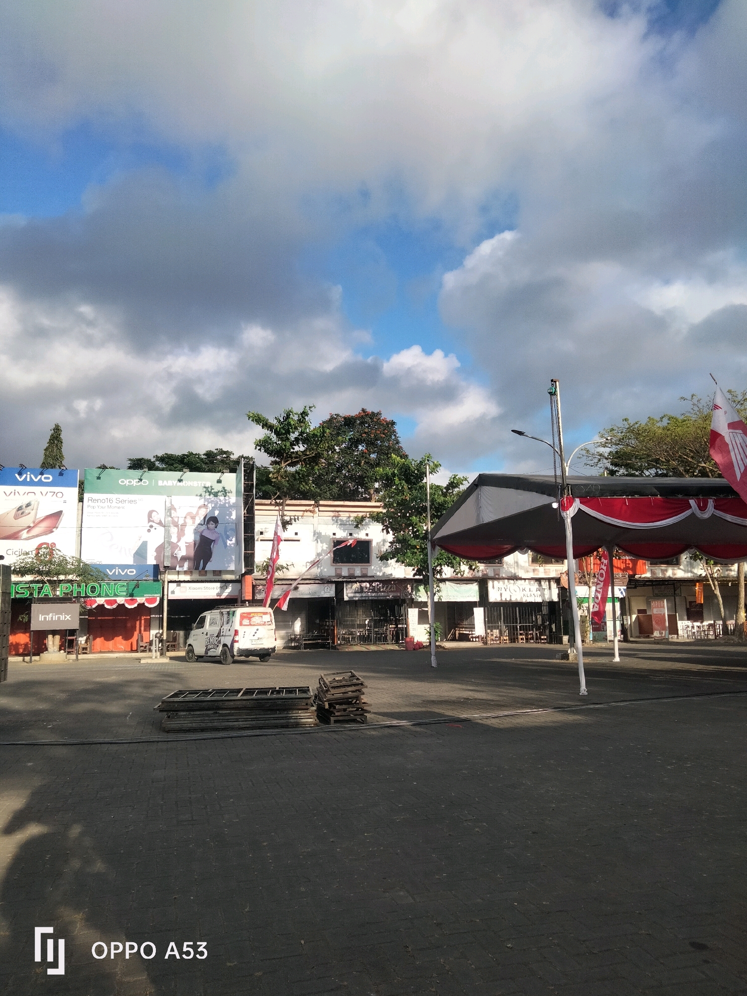 In Kelutan, Indonesia, a street scene unfolds under a partially cloudy sky, with a bright sun casting shadows. The foreground is dominated by dark, brick-paved ground, with a stack of metal frames and wooden planks on the left side. In the mid-ground, a row of storefronts lines the street, adorned with various billboards advertising brands like Vivo, Oppo, and Infinix. A white van is parked in front of one of the shops. A large, tent-like structure with red and white fabric drapes stands prominently in the mid-ground, supported by white poles. Behind the shops, lush green trees and some flowering trees add to the urban landscape. The overall impression is one of a commercial area, possibly a market or a shopping street, captured during daylight hours.