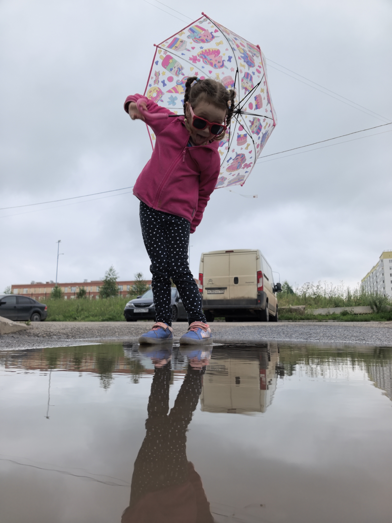 In this low-angle shot taken in Frolý, Russia, a young girl stands on the edge of a large puddle, holding a clear umbrella adorned with colorful cartoon characters. She is wearing a bright pink fleece jacket, black leggings with white polka dots, and blue sneakers. Red sunglasses are perched on her nose, and her tongue is sticking out slightly as she playfully tilts the umbrella.
The overcast sky suggests it is either a cloudy day or evening. In the background, a beige van is parked on a road, with other cars and buildings visible further away. The scene appears to be in an urban or semi-urban area, possibly a parking lot or street with a large puddle formed, perhaps from recent rain. The reflection of the girl and the surrounding environment is clearly visible in the water, adding depth to the image. The overall mood is one of childhood joy and carefree play, despite the gloomy weather. There is visible text on the license plate of the van: "X382XE 199".