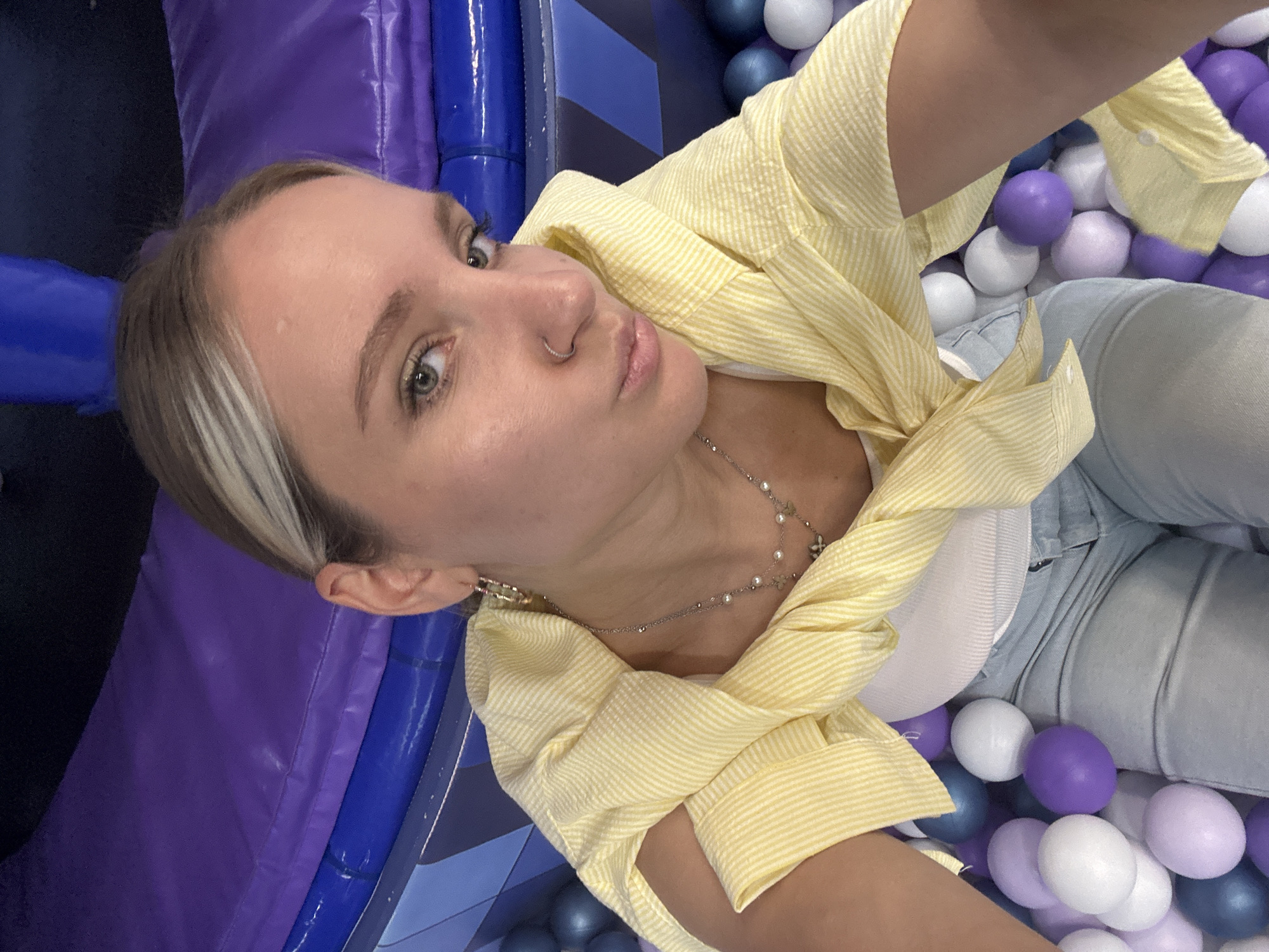 In Saint Petersburg, Russia, a young woman with blonde hair, featuring a blonde streak, poses for a selfie in a ball pit. She is wearing a white tank top, a yellow striped button-up shirt casually draped over her shoulders, and light blue jeans. She accessorizes with hoop earrings and a delicate silver necklace with pearl and floral charms. A silver nose ring adorns her nose. The background consists of a purple and blue inflatable structure, possibly part of an entertainment center, and a multitude of multi-colored balls—white, purple, and blue—filling the ball pit. The lighting is bright, suggesting an indoor environment.