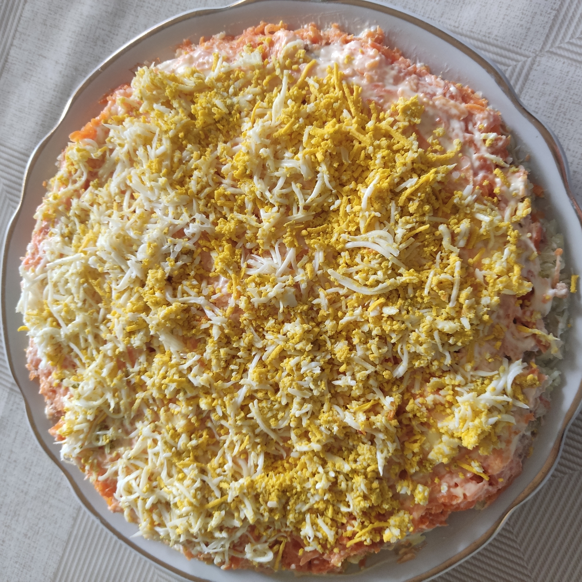 A close-up, overhead shot shows a circular salad on a white plate with a gold rim. The salad is layered, with a base that appears to be composed of shredded carrots and a creamy dressing. The top layer consists of finely grated hard-boiled eggs, with distinct sections of bright yellow egg yolk and white egg white. The salad is piled high in the center, creating a slightly domed shape. The plate rests on a textured, light-colored tablecloth. The lighting suggests it is daytime, possibly indoors, with natural light illuminating the food. There are no people visible in the image, and the focus is entirely on the presentation of the salad.