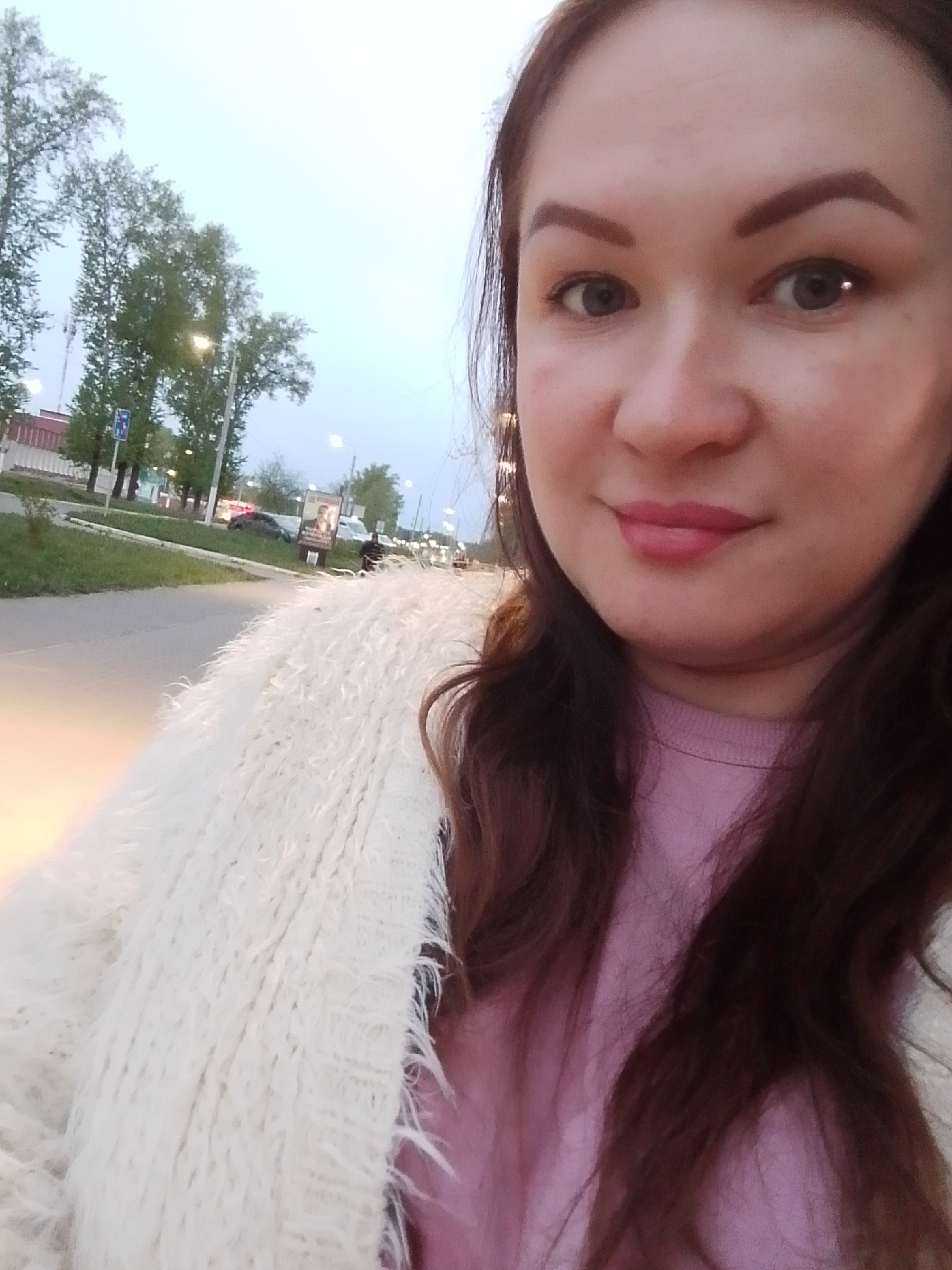 A selfie taken at dusk shows a woman with brown hair and eyes, wearing a pink sweater and a white, shaggy cardigan. She is positioned on a roadside in what appears to be the city of Chaikovsky, Russia, judging by the Cyrillic text on a billboard behind her, which is partially visible. Trees and streetlights line the road, and cars are parked in the background. There is a sidewalk next to the road, and it seems someone is walking or cycling in the distance. The lighting suggests it is either early morning or late evening. The woman is looking directly at the camera with a slight smile.