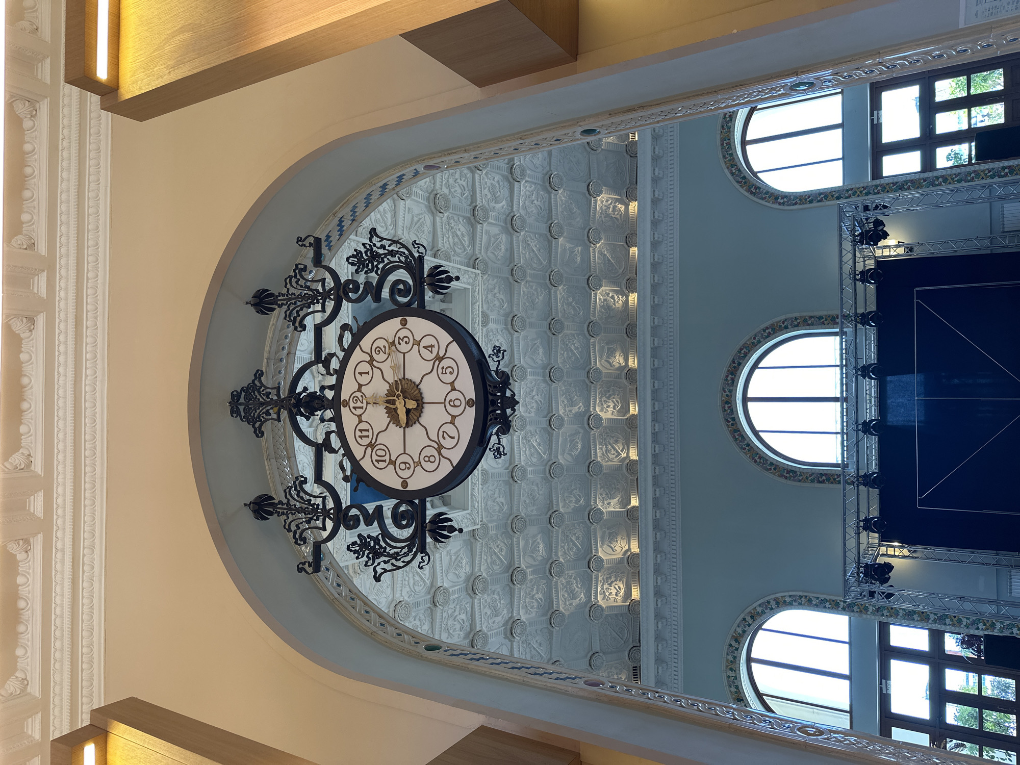 The image captures an upward perspective inside a grand and ornate interior space, likely a hall or atrium of a large public building. Dominating the central area is a large, elaborate black wrought-iron clock suspended from a decorative archway. The clock features a white face with black Arabic numerals (1 through 8, 10, 11, and 12 are clearly visible) and golden hands, indicating the time is approximately 10:20 to 10:25.
The architectural style is grand, with high ceilings adorned with intricate, repetitive patterned panels, possibly depicting heraldic shields or elaborate motifs, in shades of white and grey. Ornate moldings, cornices, and decorative pillars are visible along the arches and walls. The walls are painted in a light blue-grey, complementing the cream-colored archways and decorative elements.
Natural light streams in from multiple windows on the right side of the frame, including several arched and rectangular panes. Below these windows, a modern metal truss structure equipped with spotlights and a large dark screen (likely for projection or display) indicates a stage setup for an event or performance. Wooden ceiling beams with integrated linear lighting fixtures are visible at the periphery of the image. The scene suggests a blend of historical grandeur with contemporary event infrastructure. No people are visible. The visible text is limited to the numerals on the clock face. The overall impression is one of a majestic, well-maintained space, possibly hinting at a significant public building in Sochi, Russia.