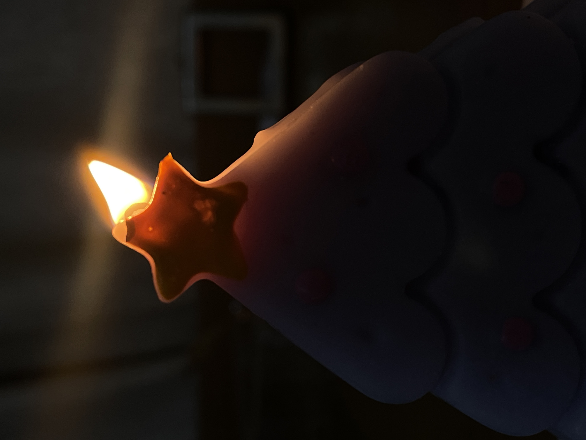 The image presents a close-up view of a burning candle, stylized to resemble a Christmas tree. The bright, warm yellow and orange flame is prominently displayed at the tip of the candle. Directly below the flame, a distinct star-shaped detail, appearing to be integrated into the candle's design and possibly slightly melted, is visible. The main body of the candle shows a layered, scalloped texture, consistent with the shape of a conifer, and is a light, somewhat translucent color. Small, reddish, circular elements are discernible on its surface, suggesting decorative accents. The background is dark and out of focus, indicating a low-light environment, with a faint, lighter rectangular shape vaguely visible in the distance. The scene captures the candle actively burning, serving as the sole significant light source, implying a nighttime setting or a dimly lit interior in Denpasar, Indonesia. No individuals or visible text are present.