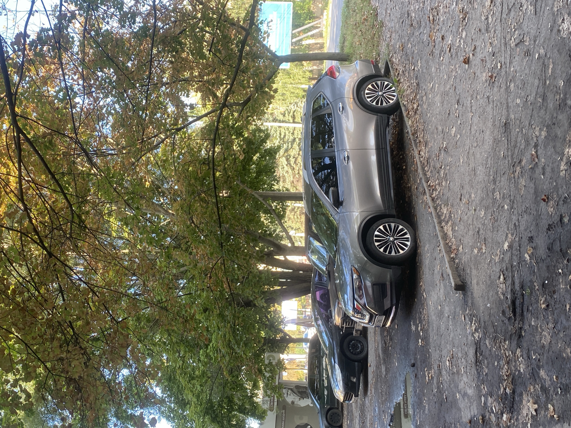 The image depicts a row of parked cars on a wet, leaf-strewn asphalt lot in Poltava, Ukraine. The cars are situated beneath a canopy of mature trees with foliage exhibiting autumnal colors, indicating the season is likely autumn.
In the foreground, a silver Nissan Rogue SUV is parked, facing towards the right of the frame. Its front is partially visible, showing the headlight and grille. The car's wheels are black with silver accents. The lot surface is uneven and appears muddy in places, with puddles of water visible. Fallen leaves are scattered across the ground.
Behind the Nissan, a dark-colored Peugeot 406 sedan is parked, also facing right. Its front end is more visible, revealing a dented bumper and black alloy wheels. A dark-colored sedan, possibly a BMW, is parked further back on the left, with its rear obscured by the Peugeot. Another car, a dark sedan with a purple tint on its windows, is partially visible between the Peugeot and the Nissan.
In the background, a building with large windows is visible behind the parked cars. Further back, trees line a roadside, and a large blue billboard is mounted on poles. The sky is visible through the tree branches, appearing partly cloudy with patches of blue. The lighting suggests it is daytime, possibly late afternoon, with sunlight filtering through the leaves. No people are visible in the image. The scene conveys a sense of everyday urban life in Poltava, with parked vehicles and autumnal weather conditions.
