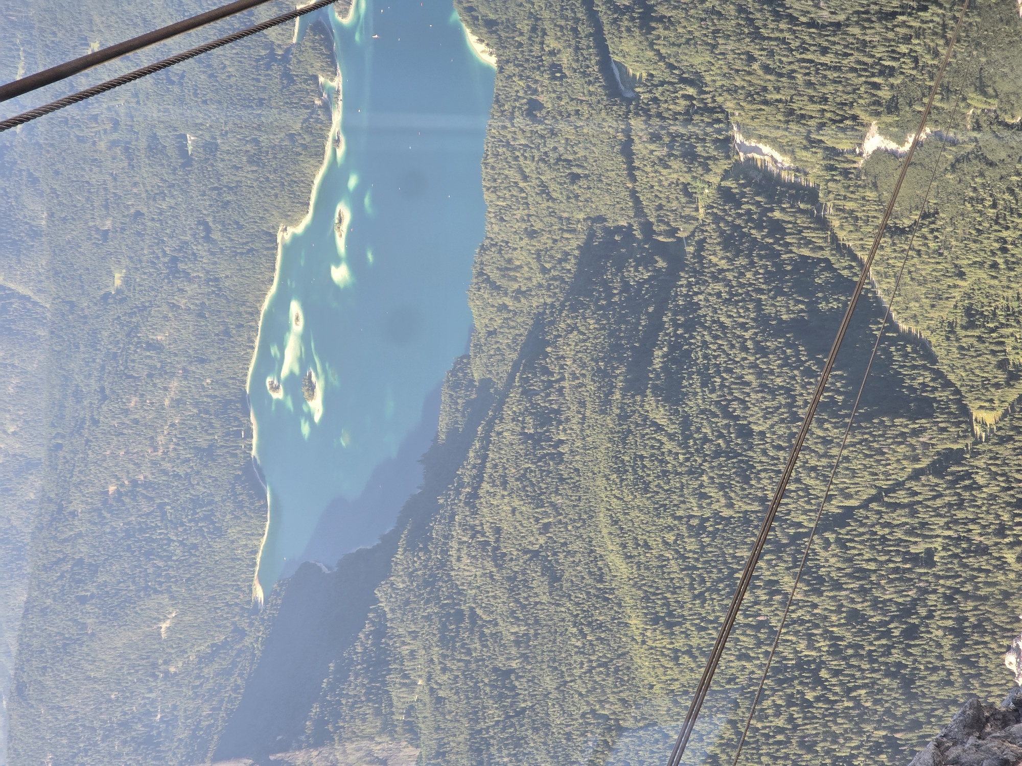The image shows an aerial view of a lake and a forested mountain landscape. In the foreground, there are cable car cables. The lake is a vibrant turquoise color and has several small islands. The surrounding mountains are densely covered with evergreen trees. The scene appears to be a natural landscape, possibly in the Bavarian Alps, near Garmisch-Partenkirchen, Germany, given the mention of that location. There are no people visible in the image, but there are a few boats on the lake and some structures or clearings within the forest, suggesting human presence in the area. The lighting suggests it is daytime, with the sun casting shadows across the landscape.