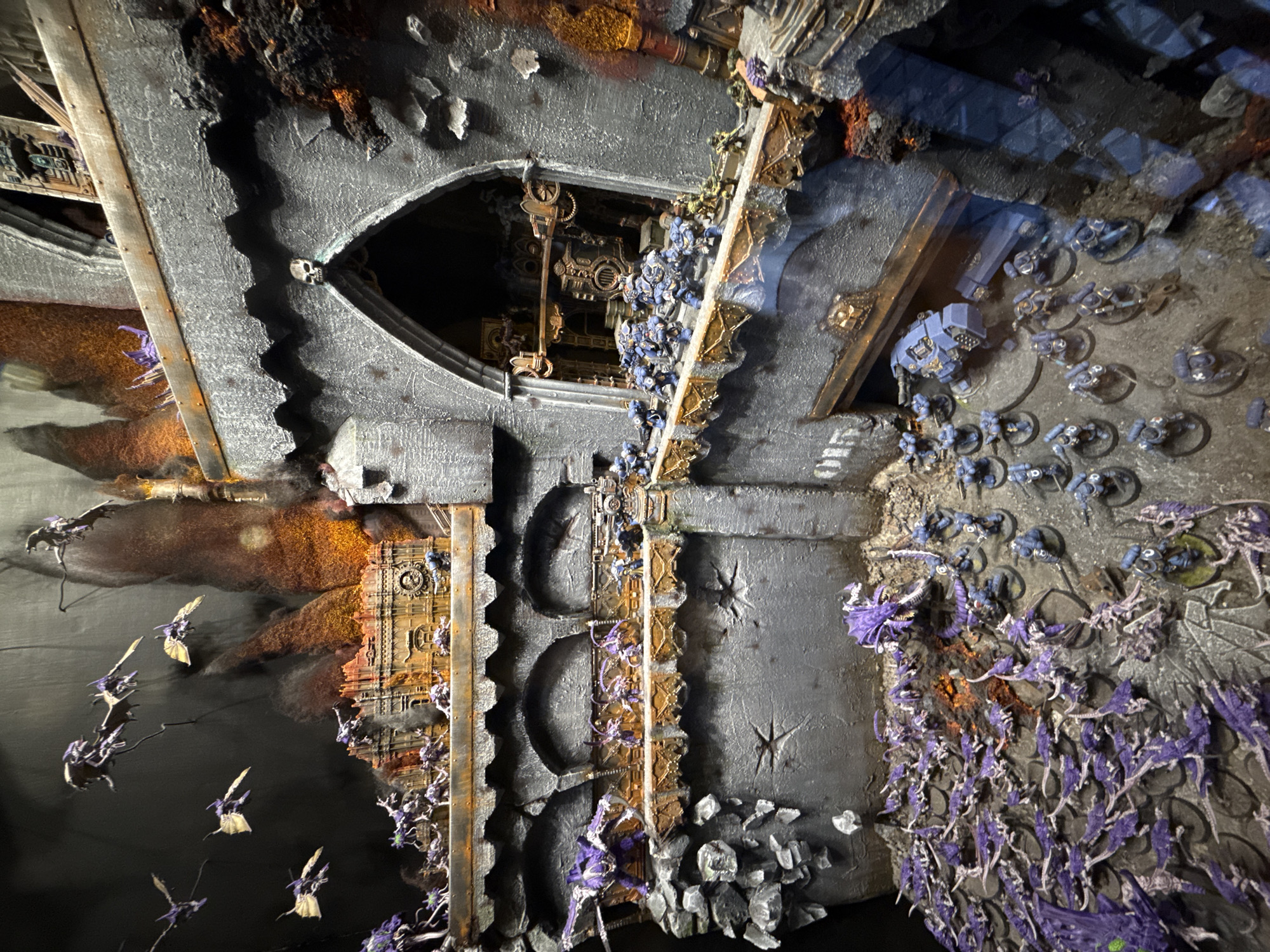 The image shows a miniature diorama of a war-torn environment. The scene features grey, heavily textured terrain with building ruins and signs of destruction. Miniature figures are scattered throughout, including groups of small, blue-armored soldiers, and winged creatures with purple accents. The diorama appears to be meticulously crafted, with attention to detail in the texture of the ruins and the arrangement of the figures. There is no indication of a specific location, and there is no connection to Barcelona, Spain.