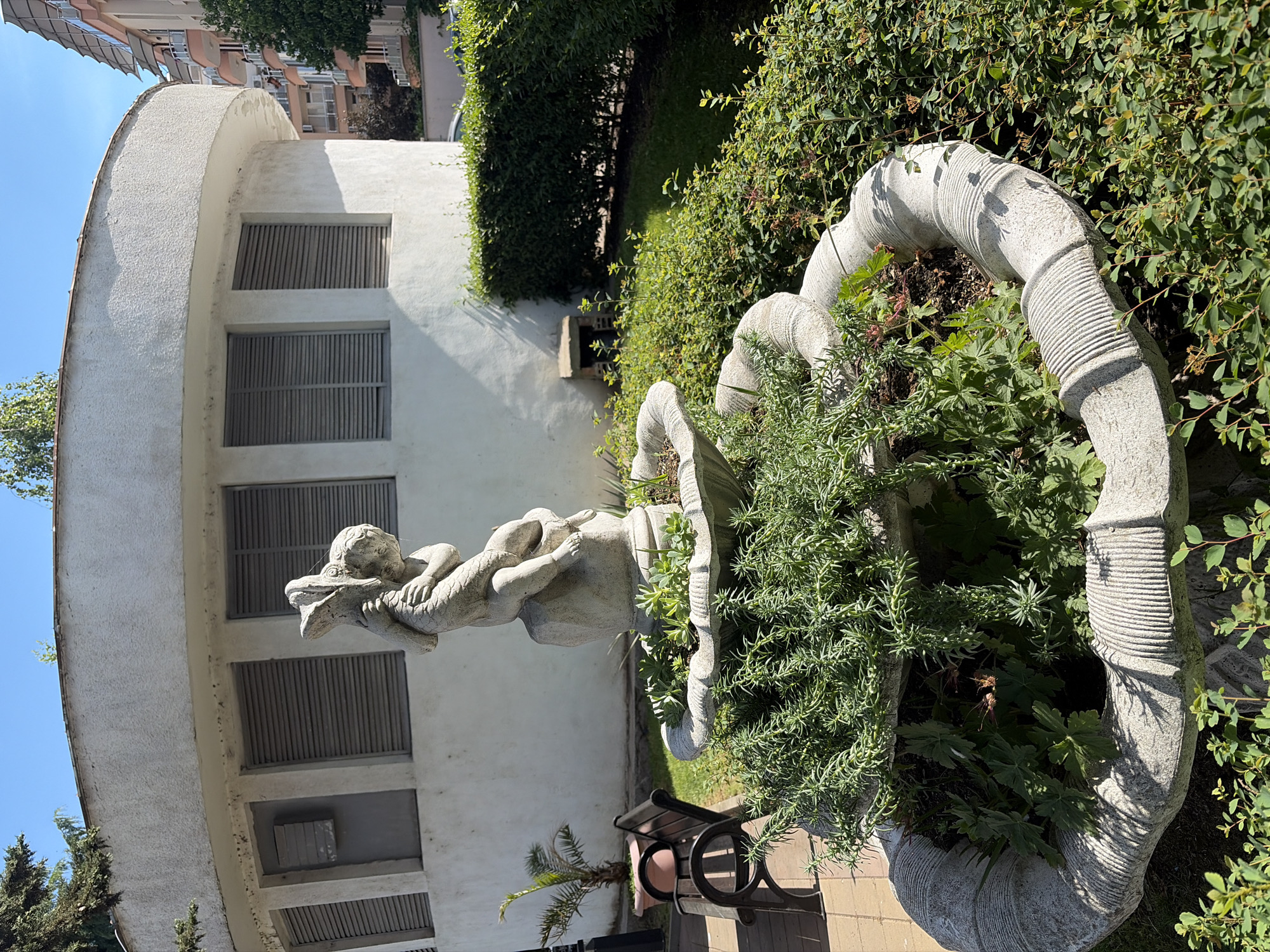 The image shows a garden scene, likely in an urban setting, with a notable statue of a child figure riding a dragon or mythical creature. The statue is placed atop a decorative, spiraling planter filled with various plants. In the background, a white, curved building with shuttered windows is visible. A bench is positioned nearby, and lush greenery surrounds the area. The overall impression is that of a well-maintained, possibly private garden. The location details are "Unknown, Unknown."
