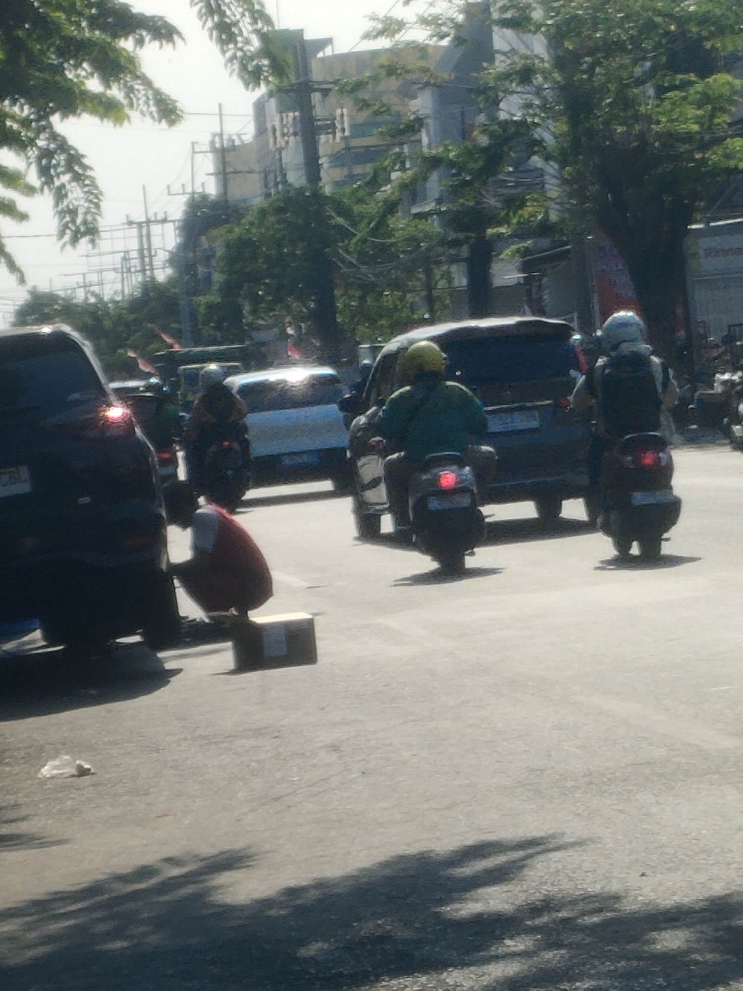 The image depicts a street scene in Surabaya, Indonesia, likely during the daytime, with traffic and pedestrians. In the foreground, a person wearing a red vest and white shirt is crouched next to a cardboard box on the asphalt road. To the left, the rear of a dark-colored SUV is visible, with its brake lights illuminated. Further into the scene, several motorcycles are present. One motorcycle has a rider wearing a yellow helmet and a green shirt. Another motorcycle has a rider with a light blue helmet. In the background, buildings and trees are visible, suggesting an urban environment. The overall impression is of a busy street with various modes of transportation and activity.