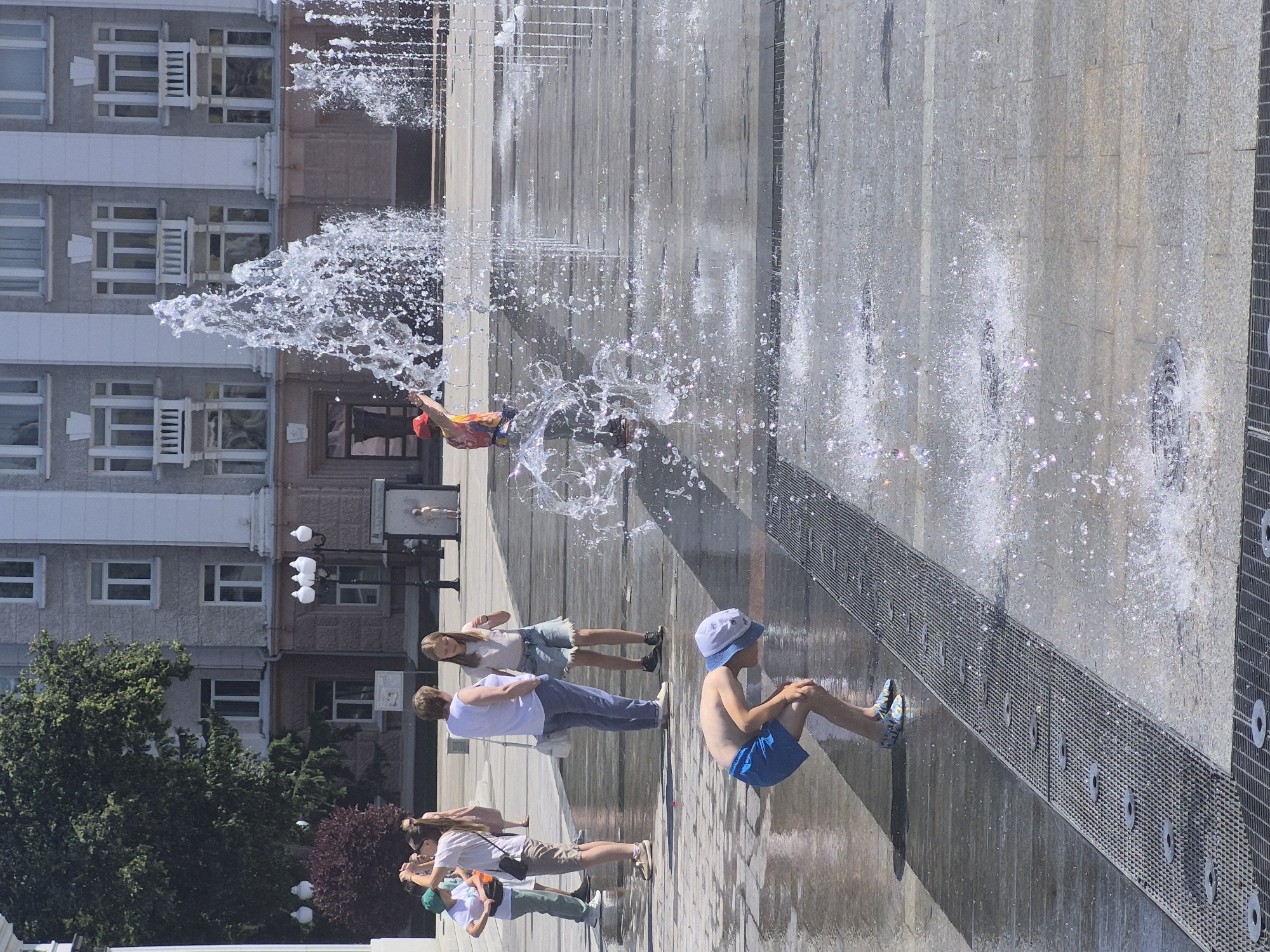 This image captures a public square with a fountain in the city of Ufa, Russia. In the foreground, a young boy wearing blue shorts and a white cap is bent over, seemingly interacting with the water jets of the fountain. He is barefoot with patterned blue and yellow shoes on his feet. Behind him, a group of people are standing in the square. On the left, three individuals, two adults and a child, are facing away from the camera. One adult, a woman in a white top and blue jeans, has a white purse slung over her shoulder. The child next to her is wearing a white t-shirt and green pants. Another woman is standing slightly to the right, facing the camera, wearing a white top and denim shorts. Further back, a woman in a white shirt and blue jeans and a young girl in a t-shirt and denim shorts are conversing. In the background, a multi-story building with light-colored walls and numerous windows is visible, suggesting an urban setting. A large green tree with dense foliage is positioned to the left of the building, and a smaller reddish-purple shrub is visible in front of it. The ground of the square is paved with stone slabs and features metal grates, indicative of a fountain system. Water from the fountain is spraying upwards and outwards, creating a dynamic and refreshing atmosphere. The sky is clear and blue, suggesting a sunny day.