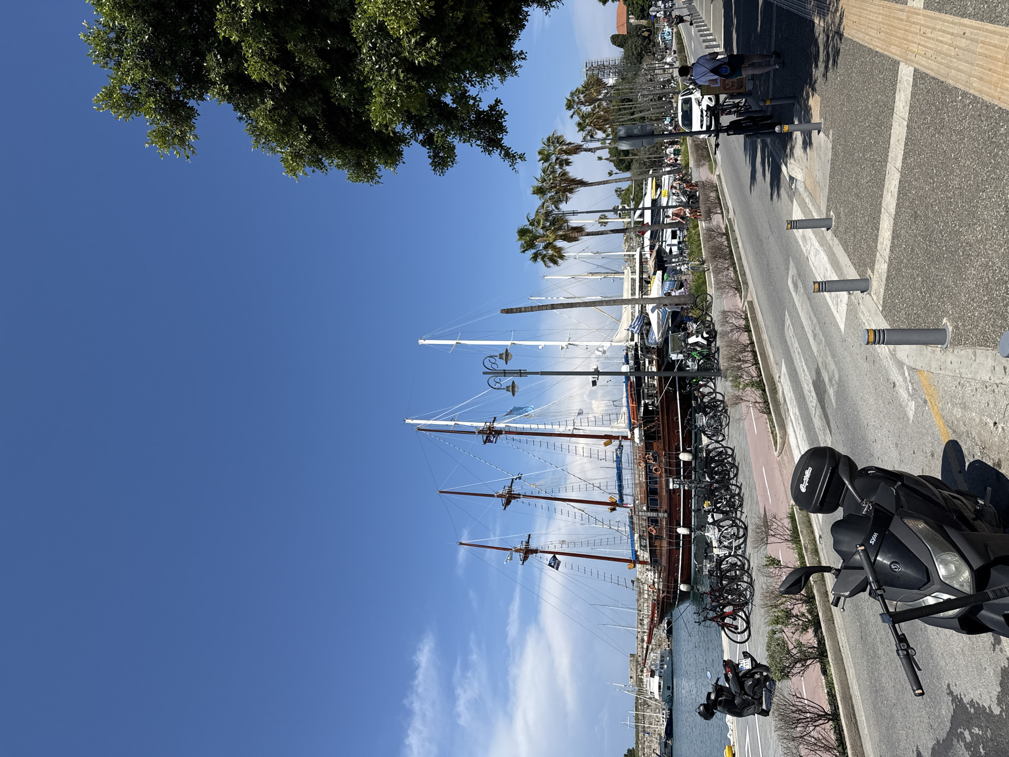 The image shows a view of Kos harbor, in the city of Kos, Greece, on a sunny day. A large, three-masted wooden ship, likely a tourist vessel or replica pirate ship, is docked alongside the harbor. There are numerous bicycles parked in a row between the ship and the road. A black scooter is parked in the foreground. The harbor and masts are adjacent to the road and pedestrian walkway. The roadway has painted pedestrian crossing, separated by gray bollards. The walkway has a textured surface. The background includes palm trees, other boats in the harbor, buildings, and other palm trees in the distance, with some visible construction scaffolding in the distance. Clear blue skies are overhead, with a few scattered clouds, and tree foliage is in the upper left corner. Some pedestrian traffic is seen, with at least 3 people visible.