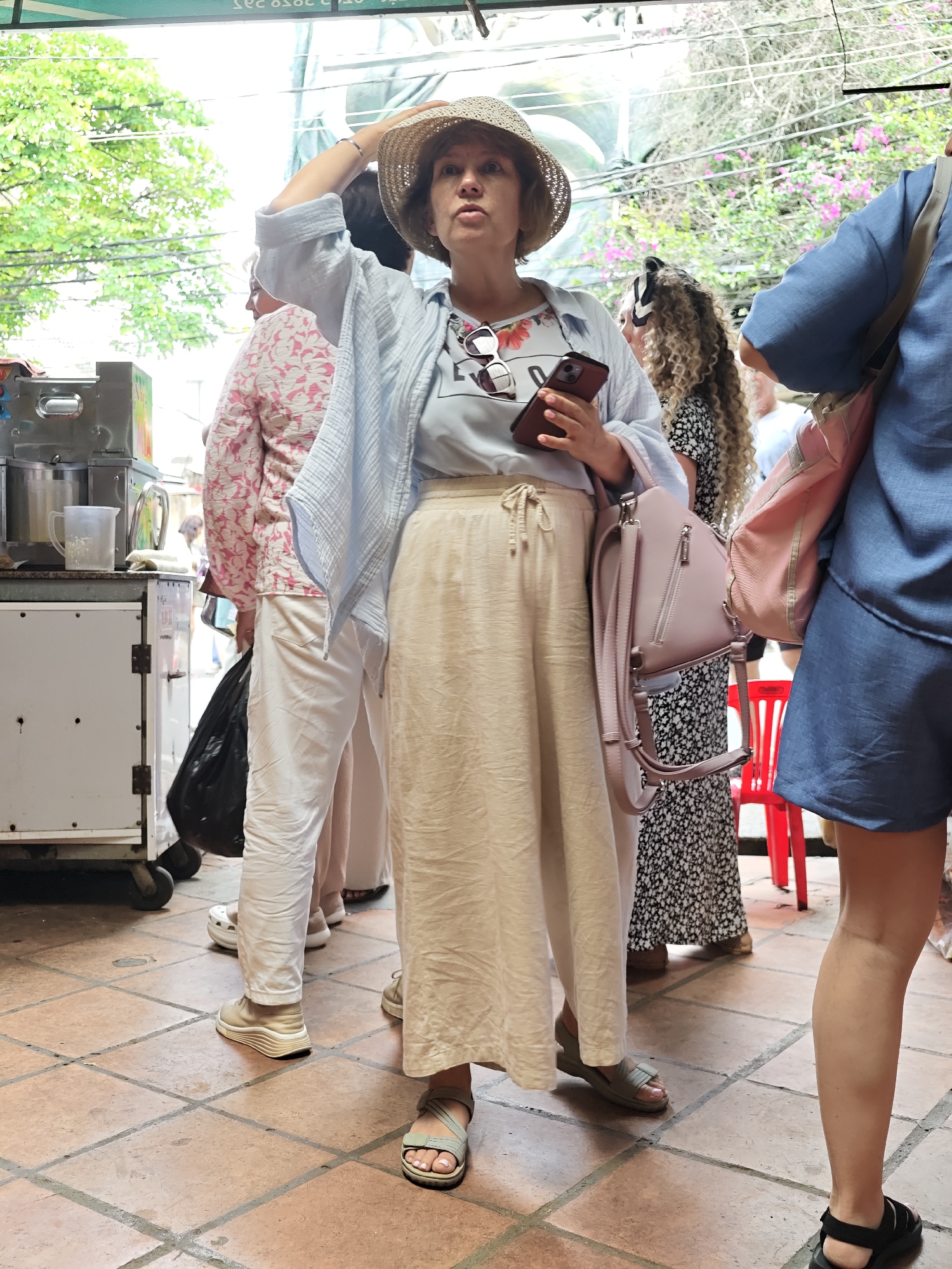 The image depicts a street scene in Da Lat, Vietnam, where a woman wearing a straw hat and a light blue shirt is holding a smartphone. She is also carrying a light pink backpack. To her left, another person is partially visible wearing a pink floral-patterned shirt and white pants. In the background, greenery and flowers can be seen, suggesting an outdoor setting. The ground is paved with terracotta tiles. The overall atmosphere appears to be casual and perhaps a tourist-oriented location given the presence of the phone and backpack.