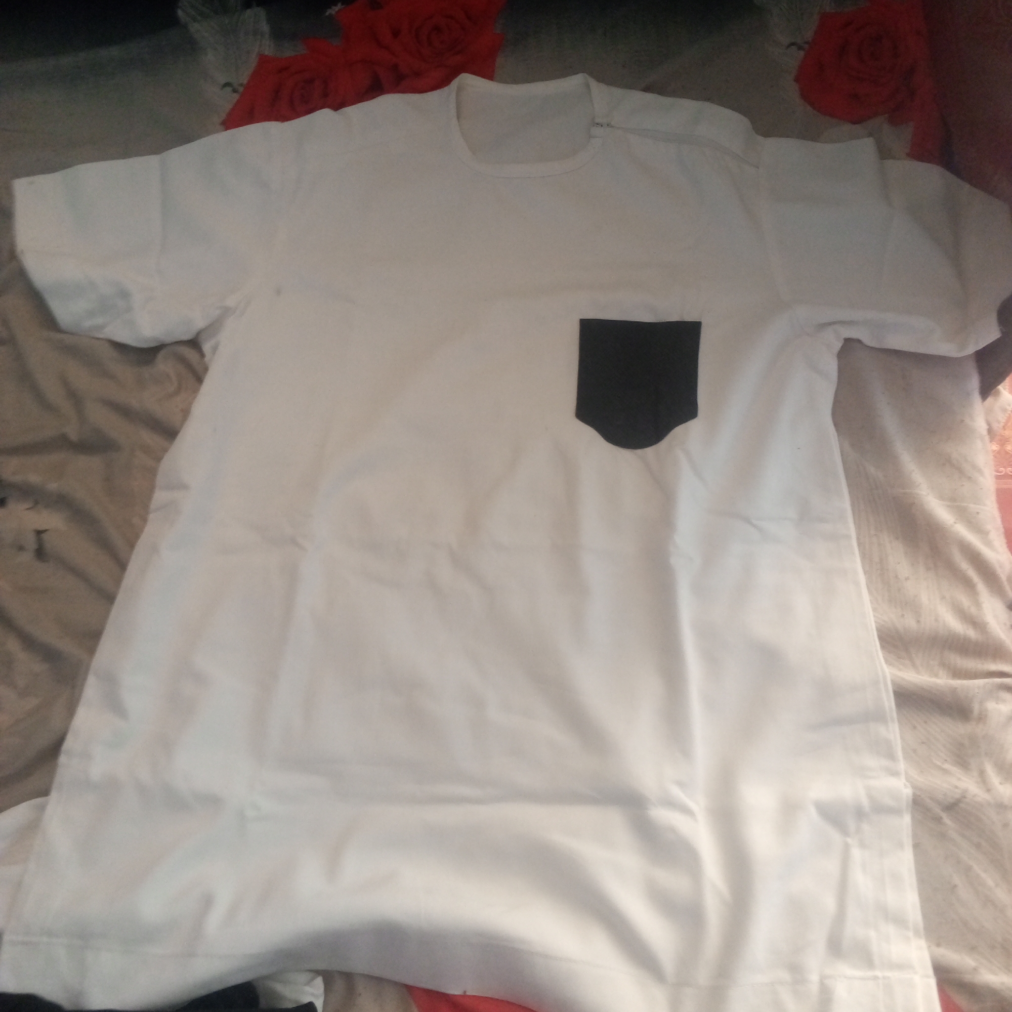 The image shows a white t-shirt with a black pocket on the left side of the chest. The t-shirt is laid flat on a surface that appears to be a bed or sofa, covered with a patterned fabric. The background is blurred and shows hints of red fabric with floral patterns, possibly pillows or a blanket. The lighting suggests an indoor setting. There is no visible indication of the city of Zinna, Nigeria, or any other specific location. The image is a close-up shot focusing on the t-shirt.