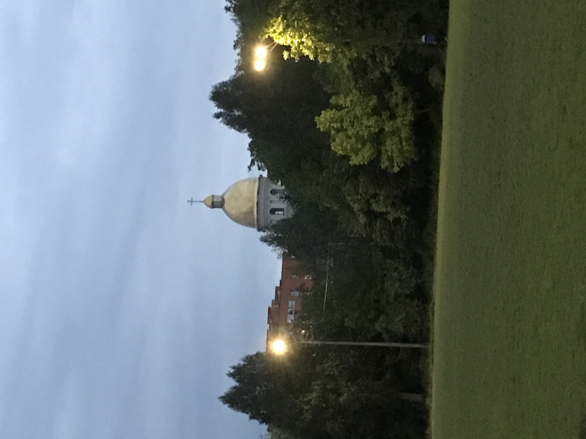 The image depicts a cityscape at dusk or dawn. A building with a golden dome and a cross is partially visible behind trees. The architecture suggests a church or cathedral. In the foreground, there's a grassy area. Street lights illuminate the scene. The image is taken in the city of Ternopil, Ukraine.