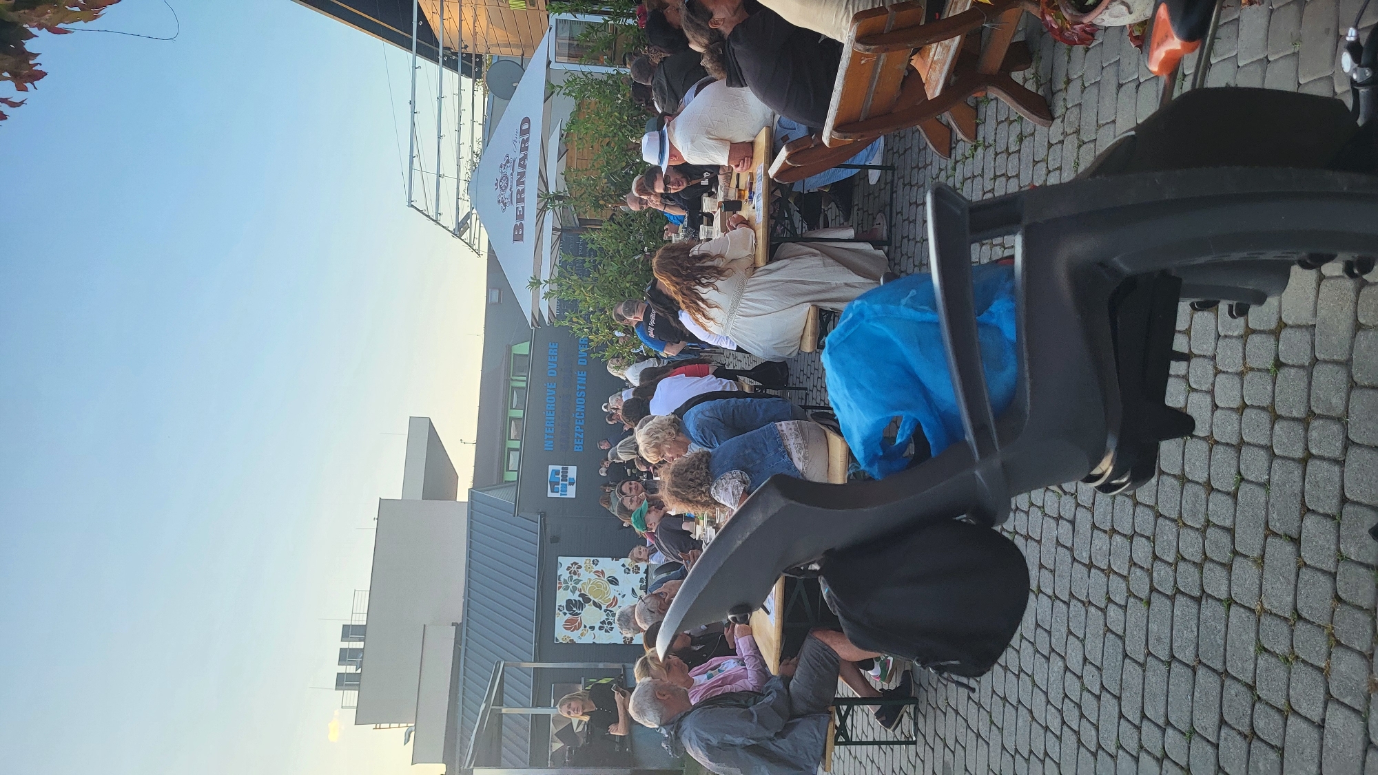 The image depicts an outdoor gathering of people sitting at picnic tables in what appears to be a brewery or beer garden, potentially in Prešov, Slovakia, indicated by the sign "BERNARD" on an umbrella. In the foreground, a dark gray child's bicycle seat is prominently featured, obscuring part of the view. Behind the seat, several people are seated at tables, some with drinks and food. The setting includes a paved ground with interlocking stones and a building with signage that reads "INTERIÉROVÉ DVERE" and "BEZPEČNOSTNÉ DVERE," suggesting a business selling interior and security doors. The sky is clear and bright, indicating daylight. The overall atmosphere seems relaxed and social.