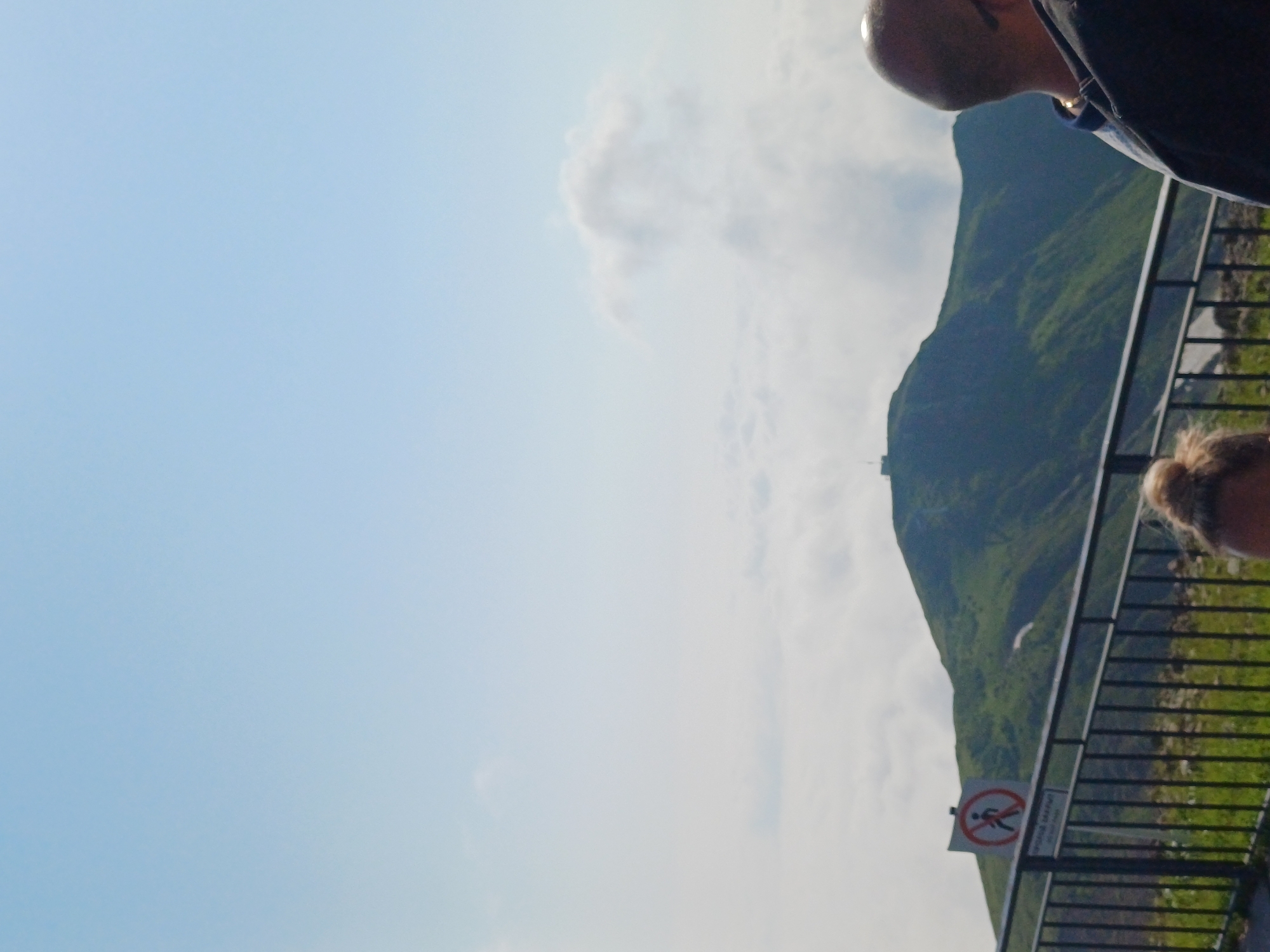 The image shows a panoramic view from a mountain peak in Estosadok, Russia. In the foreground, there is a black metal fence with a sign indicating "Do not pass" in a red circle with a diagonal line, and a person's head with blonde hair visible. To the right, a man with a bald head and sunglasses is partially visible. Behind the fence, a steep, green mountain rises, topped with a small structure. A layer of white clouds obscures the lower parts of the mountains, creating a sea of clouds effect. The sky is clear and blue. The overall scene suggests a scenic overlook on a clear day.