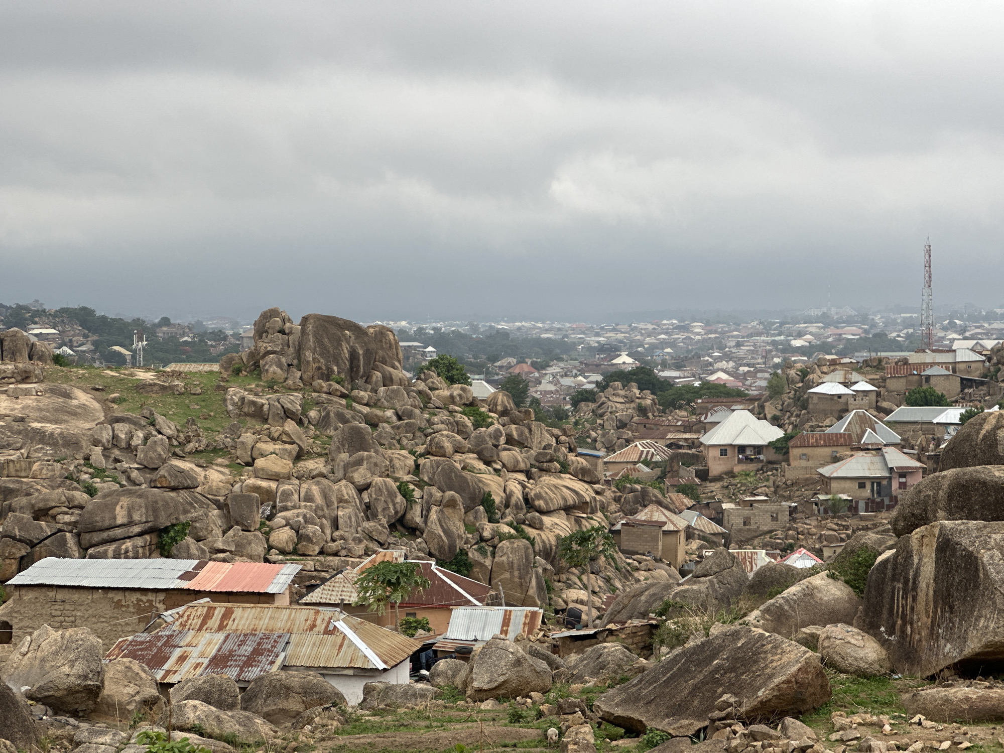 The image captures a cityscape in Jos, Nigeria, characterized by a rugged landscape interspersed with human settlement. Large, rounded gray-brown boulders dominate the foreground and middle ground, scattered among and around dwellings. These houses are simple structures with corrugated metal roofs of various colors, including silver, orange, and rusty brown. The buildings are integrated into the rocky terrain, creating a unique architectural landscape. In the background, a dense urban area stretches out under an overcast sky. A tall, red-and-white communication tower rises above the city skyline. The weather appears cloudy and potentially rainy. The scene suggests a close interaction between nature and human habitation in Jos.