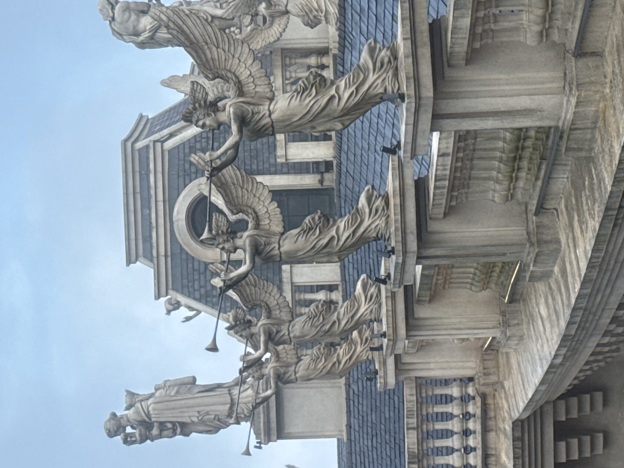 The image shows a building facade in Đà Nẵng, Vietnam, decorated with a series of angelic, winged sculptures playing trumpets. The sculptures are positioned along the roofline of the building. The building's architecture is ornate, featuring a dark blue tiled roof, arched windows, and detailed stone work. The sky is overcast.