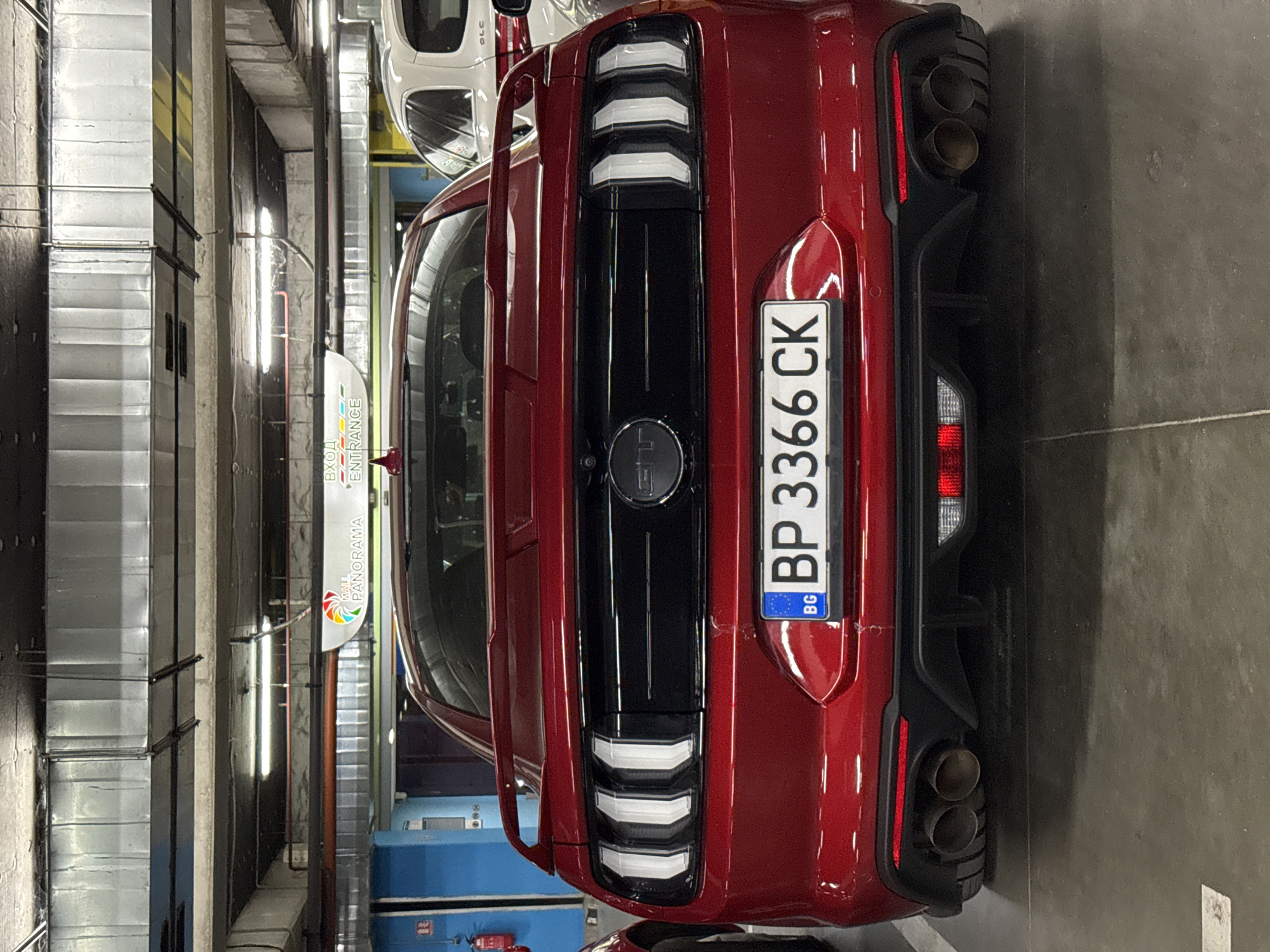 A red Ford Mustang GT is parked in a multi-level parking garage. The car faces the camera, revealing its rear end. The license plate reads "BP 3366 CK" and has a Bulgarian flag emblem. The car has a spoiler and dual exhaust pipes. In the background, a white SUV is partially visible. Overhead, fluorescent lights illuminate the concrete ceiling and ductwork of the parking garage. A sign above the car indicates "MALL PANORAMA ENTRANCE". The floor is concrete.