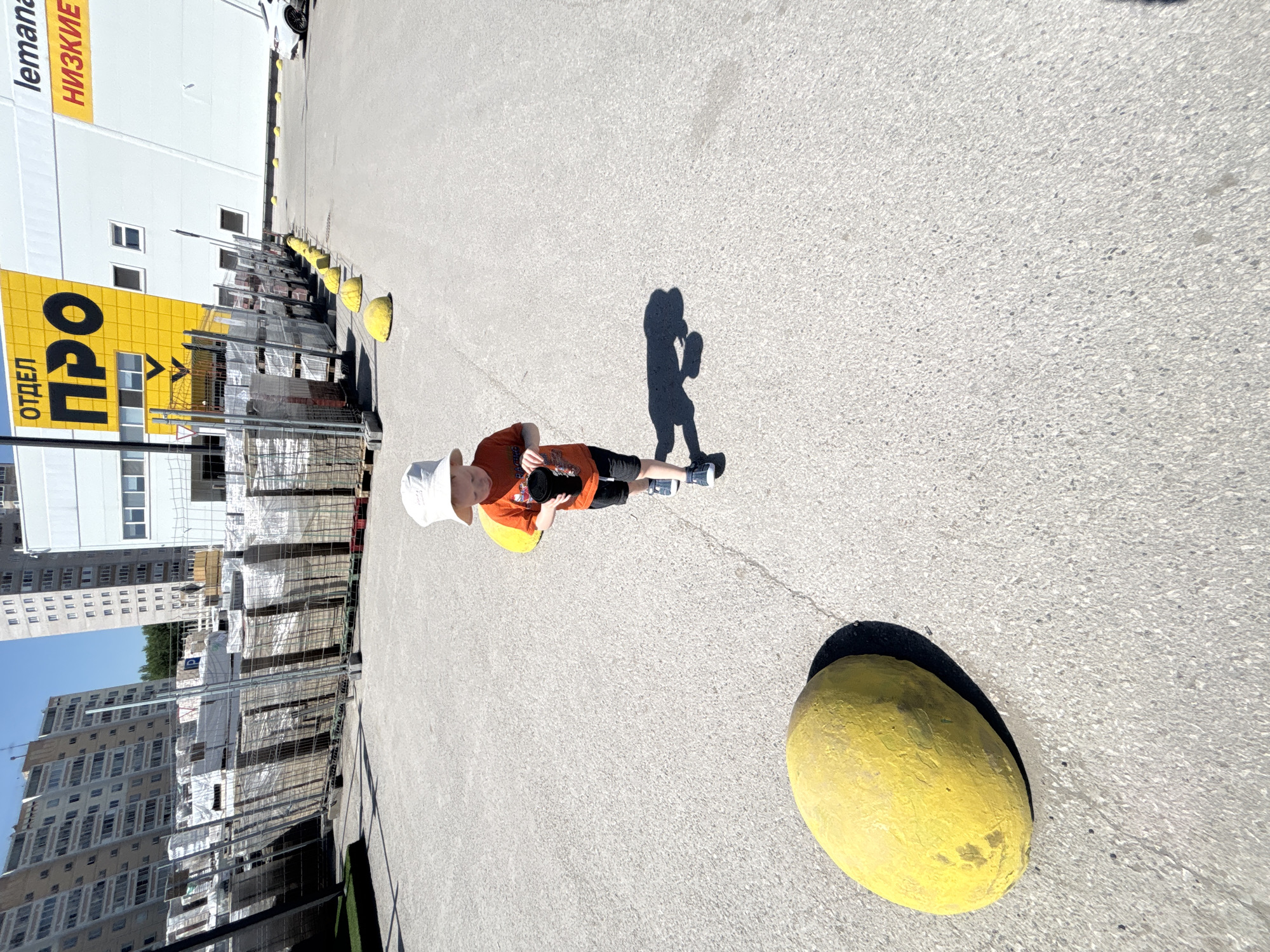 The image shows a young child standing on a paved surface in what appears to be an urban environment. The child wears a light-colored hat, an orange t-shirt, and dark shorts, and appears to be holding a camera. The ground is grey and marked with a crack. There are large yellow spherical objects in the foreground and near the child. A shadow of the child is visible. In the background, there is a building with a sign in Russian, "ОТДЕЛ ПРО" (department "PRO") and "lemana НИЗКИЕ" (lemana LOW). Behind this building, a row of metal fencing and stacked building materials can be seen. Further in the background, tall residential buildings are visible under a clear blue sky. The location is Perm, Russia, as evidenced by the Russian text on the sign.