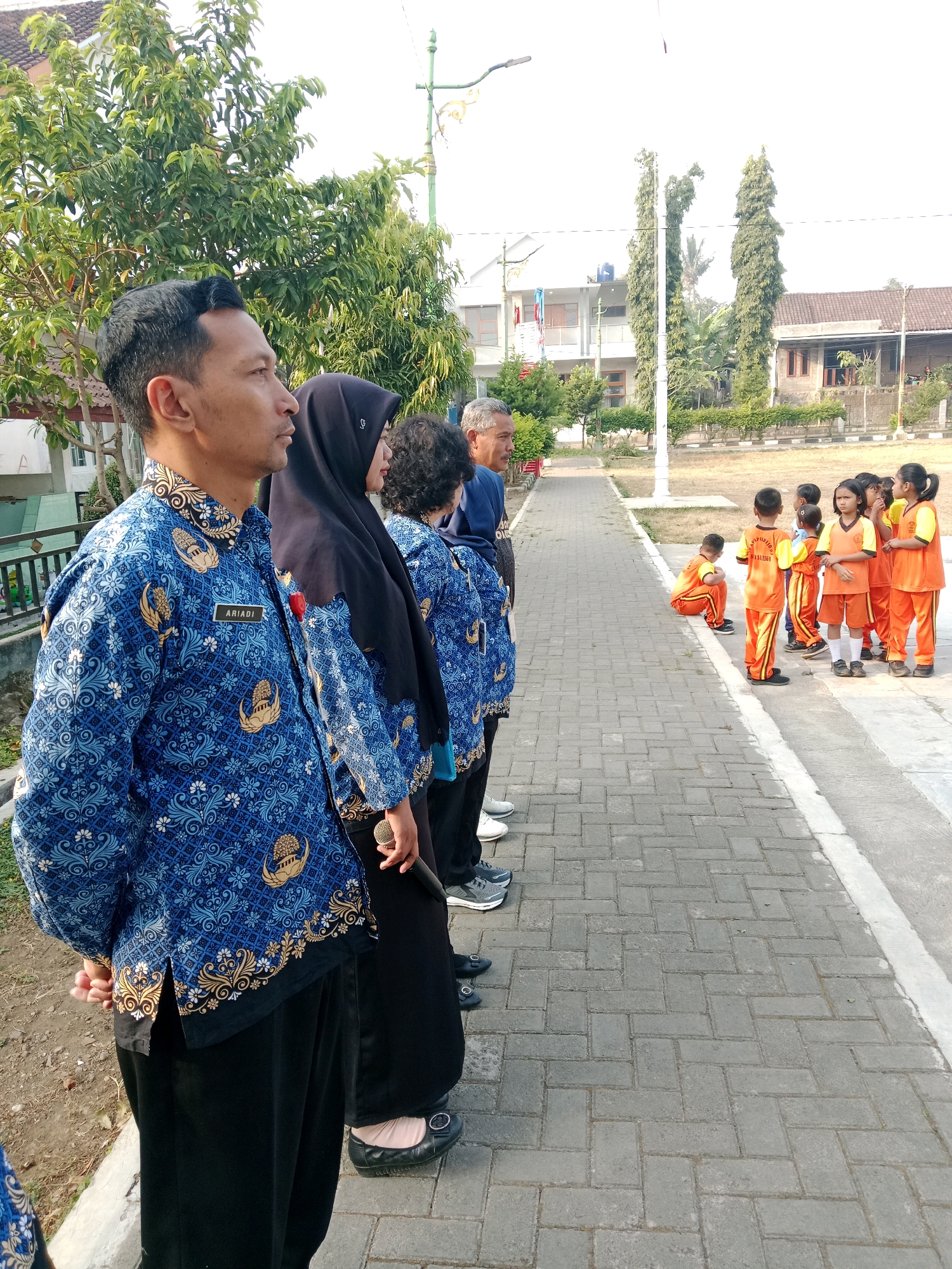 In Salatiga, Indonesia, an outdoor scene unfolds with a group of adults standing in formation, facing a group of young children. The adults, dressed in matching blue batik shirts with intricate patterns and gold accents, appear to be school staff or officials. One man in the foreground, identified by a nametag as "ARIADI," wears his shirt buttoned up and has his hands clasped behind his back. Beside him, a woman is seen wearing a black hijab.
The children, on the other hand, are dressed in vibrant orange and yellow uniforms, suggesting they are students. They are gathered to the right of the adults, some standing and others one child crouched on the ground. The setting appears to be a paved walkway, possibly within a school or community area, with a grassy field and buildings visible in the background. Tall, slender trees and smaller bushes add to the natural element of the environment. The overall atmosphere suggests a formal gathering or event, possibly a school assembly or a welcoming ceremony.