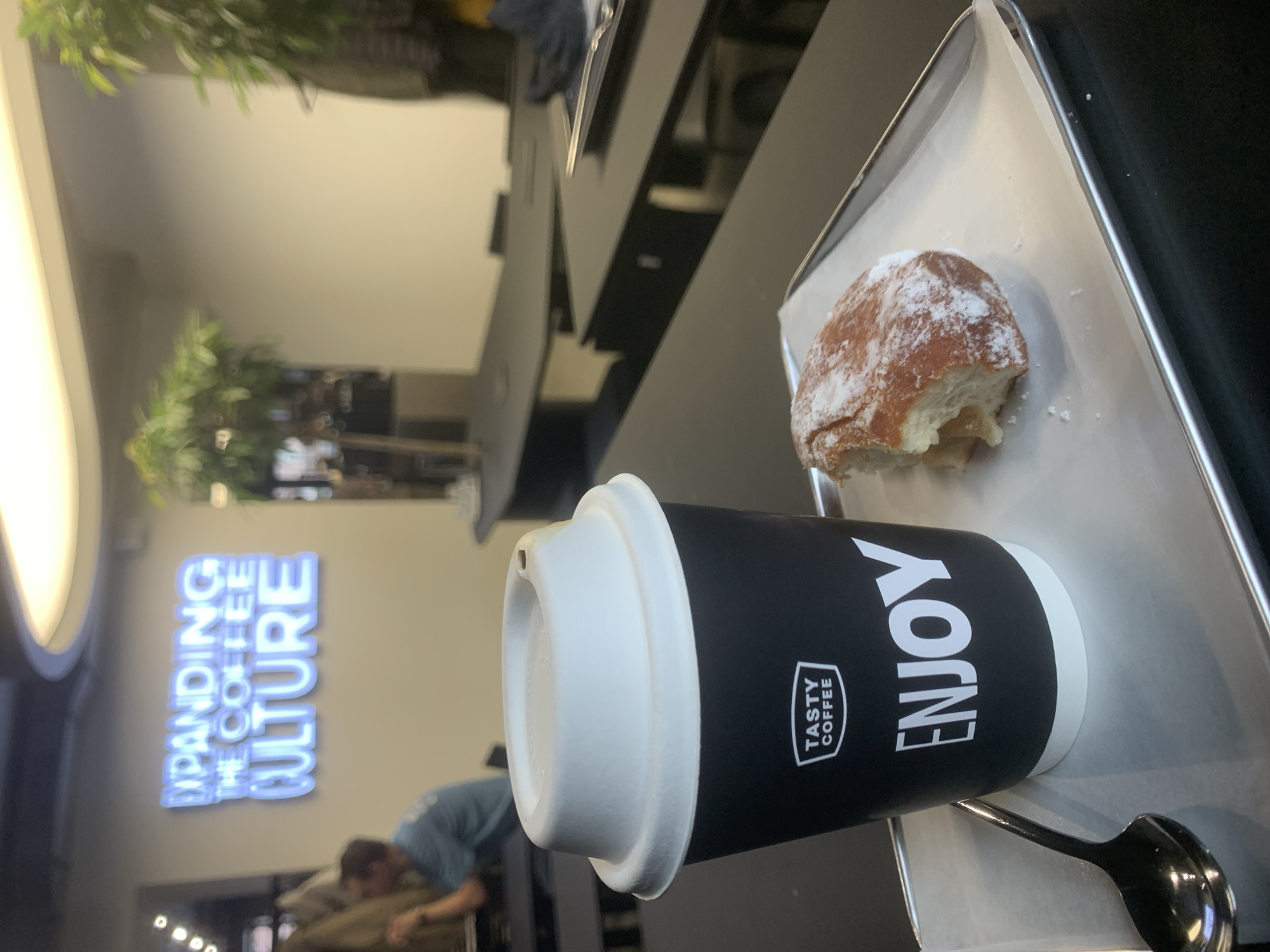 A cup of Tasty Coffee brand coffee and a half-eaten donut are on a silver tray with a spoon on a black table. The cup is black with a white lid. "Enjoy" is printed vertically on the side of the cup. In the background, a neon sign on a wall reads "Expanding the Coffee Culture" and other patrons are milling about. The scene is likely inside a Tasty Coffee shop location in Izhevsk, Russia.