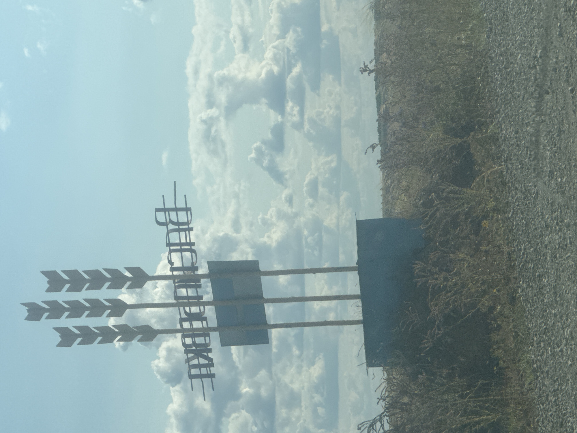 A rural signpost with three stylized wheat stalks rises against a cloudy blue sky. Beneath the wheat stalks, a metal sign is mounted on three poles, with a dark blue base. The sign features white, blocky lettering that reads "ВАСИЛЬЕВКА" (Vasilyevka), indicating the town. The sign is situated in an open field with dry grass and sparse vegetation in the foreground and background. The ground is covered with gravel. The scene appears to be captured from a moving vehicle.