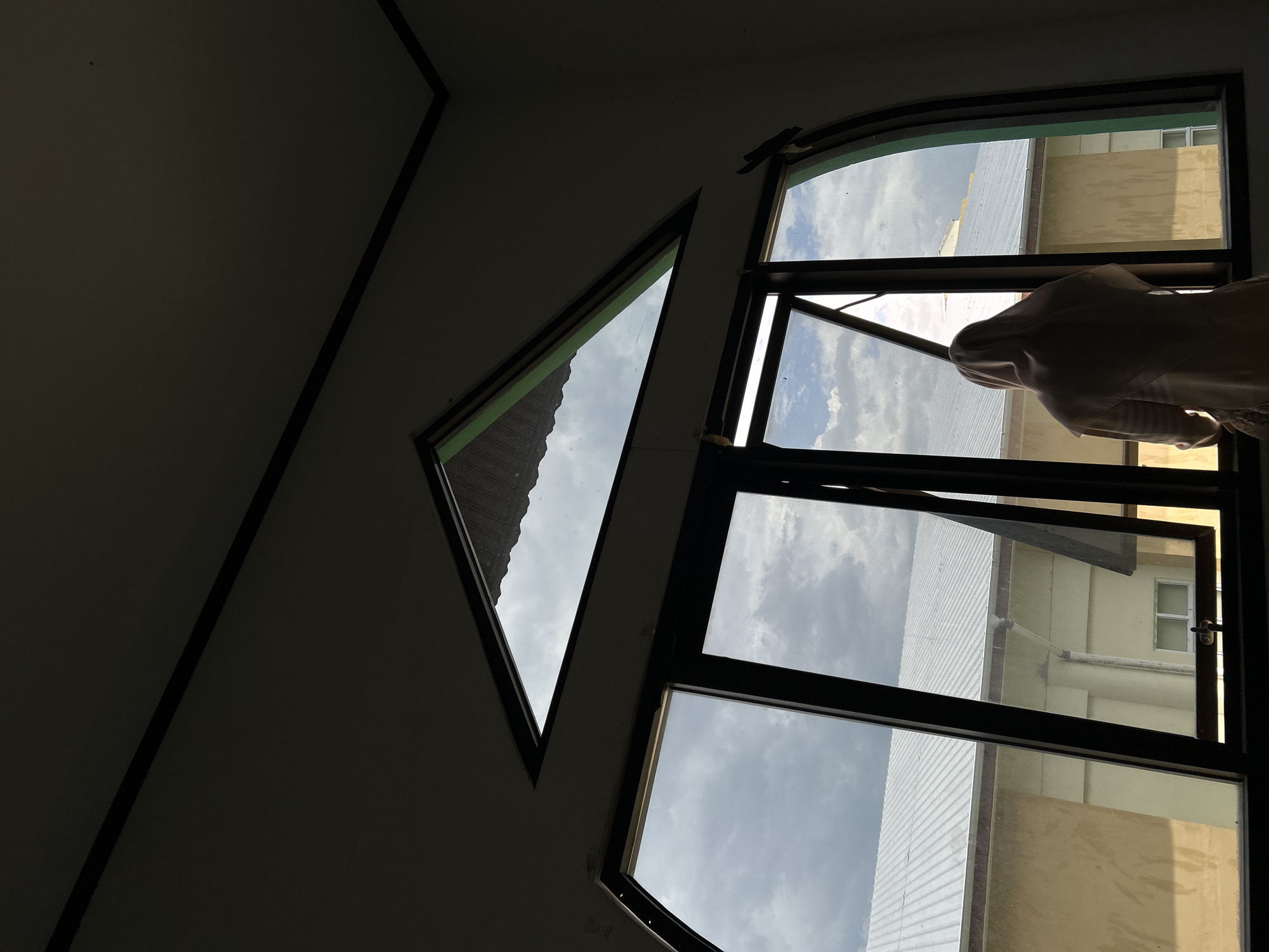 The image captures an indoor view looking out through windows during daytime in Reuleut Barat, Indonesia. The interior, consisting of a dark wall and ceiling, is dimly lit, creating a stark contrast with the bright, cloud-dotted sky and the exterior world visible through the windows.
To the right, a large rectangular window with dark frames is featured, with one pane slightly ajar. A person is partially visible, seen from the back, leaning out or looking through this window. They are wearing a light-colored garment that covers their head and upper body, possibly a hijab. To the left of this main window, a smaller, triangular window, also with dark framing, provides an additional glimpse of the outside.
Beyond the windows, parts of adjacent buildings are visible under the bright, cloudy sky. These structures feature corrugated metal roofs and light-colored walls, indicating a typical urban or semi-urban environment. The time of day is clearly daytime, given the natural light and visible clouds. No text is visible in the image.