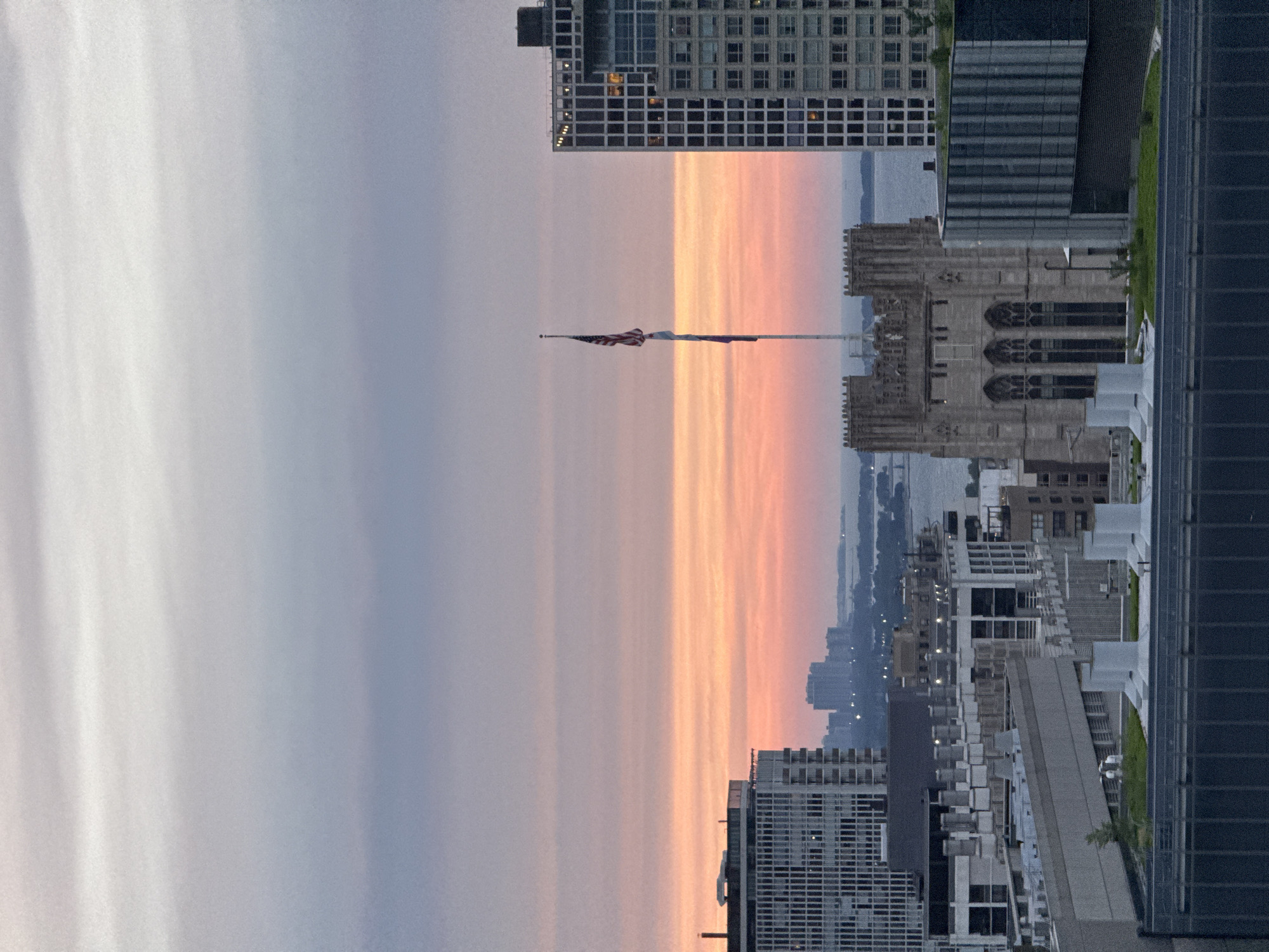 This is a high-angle, wide shot of a city skyline at dusk, likely Chicago, given the architectural style and the presence of a body of water in the background, which appears to be Lake Michigan. The sky displays a gradient of colors, transitioning from a pale gray at the top to soft pink and orange hues near the horizon, indicative of sunset.
Dominating the mid-ground is a Gothic-style building with intricate stone carvings and arched windows, possibly a university building or a historic church. To its right, a tall, modern skyscraper with a grid-like facade of windows pierces the sky, with a few illuminated windows suggesting occupancy. A flagpole with the American flag is prominently positioned between these two structures.
In the foreground, several rooftops of modern buildings are visible, featuring concrete structures, ventilation systems, and some greenery. The distant background shows a hazy silhouette of more urban development against the water. The overall atmosphere is serene and urban, capturing the transition from day to night in a metropolitan setting. No people are discernible in the image.