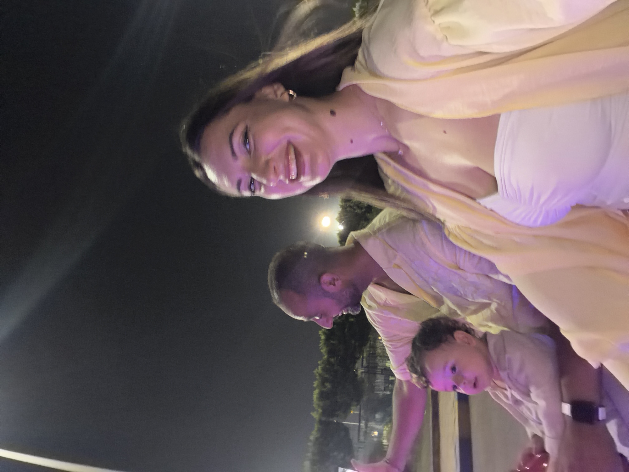 In a nighttime scene in the city of 6th of October, Egypt, a family is captured in a selfie. A man, with short hair and a beard, is visible in the background, holding a phone to take the picture and smiling. In the foreground, a woman with long brown hair smiles broadly, her head tilted slightly. She is wearing a white strapless top and a light yellow open cardigan. A young child, with curly hair, sits on someone's lap in the center of the image, looking to the left. The background shows a dimly lit urban environment with trees and streetlights. The lighting is predominantly from artificial sources, casting a purple and yellow hue on the subjects.