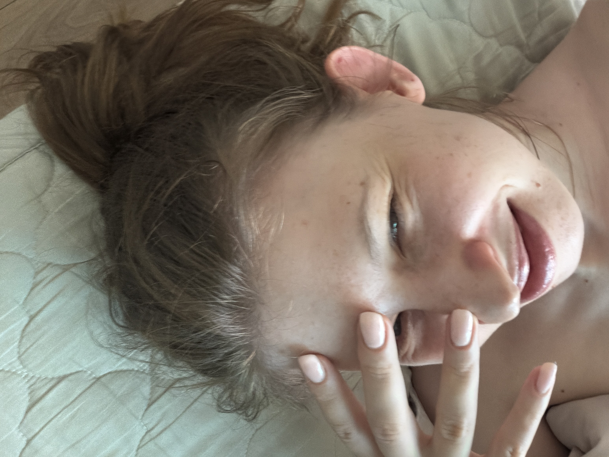 A close-up, overhead shot shows a woman lying on a light green quilted surface, possibly a bed. Her head is tilted back, and her hair is pulled up into a messy bun. She is covering one eye with her left hand, her fingers splayed, and her other fingers are visible on her cheek. Her right eye is partially visible, and she appears to be smiling or smirking. She has light skin with freckles and her nails are painted a pale pink. The background consists of the textured green surface. No specific location in Dongducheon-si, South Korea can be identified from the image.