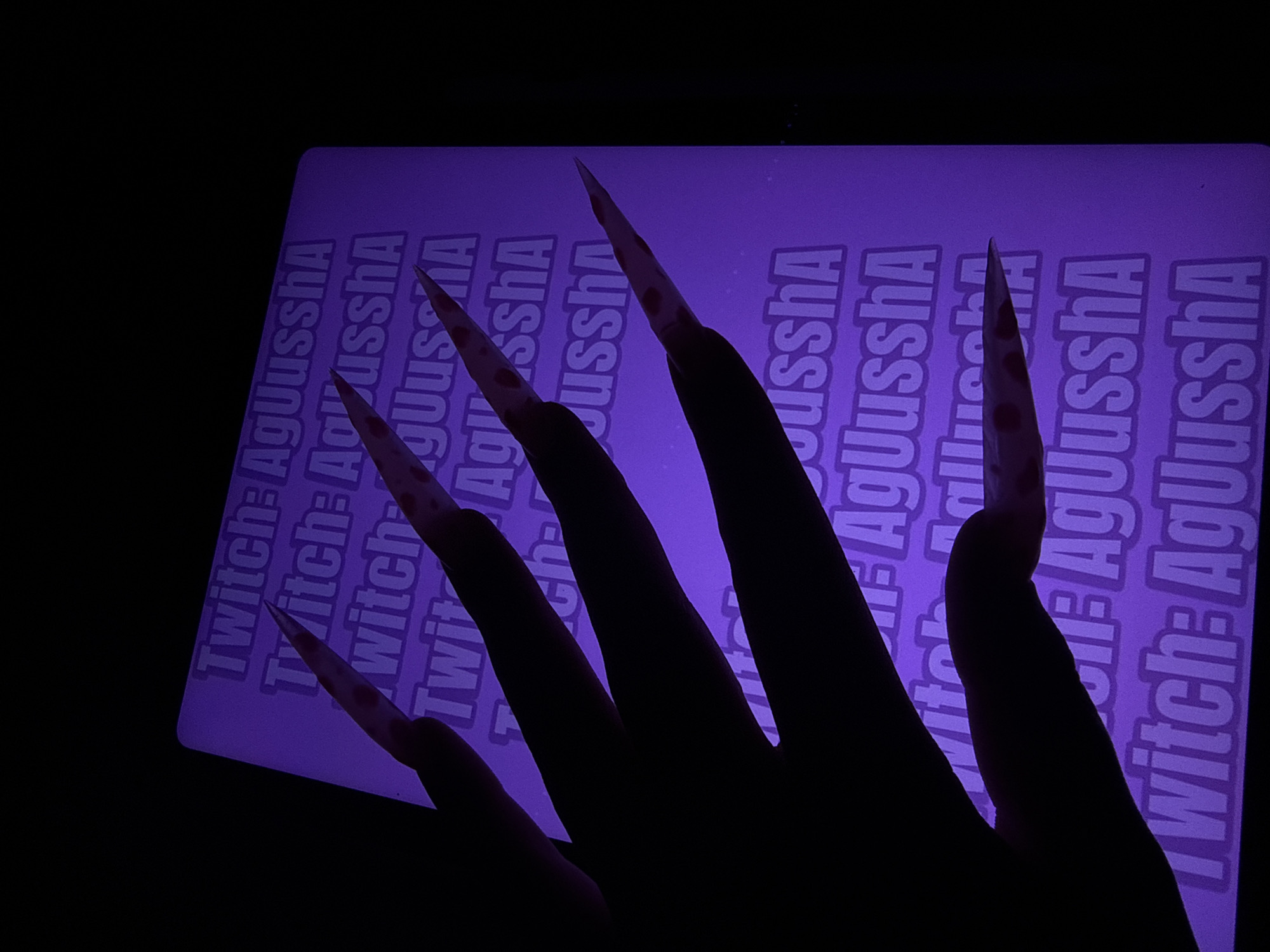 A hand with long, pointed nails adorned with dark red speckles is silhouetted against a glowing purple surface. The surface displays repeating text that reads "Twitch: AgUusshA" in a stylized font. The image appears to be taken in a dark environment, possibly a room, with the purple glow emanating from the surface. No specific location in Pudomyagi, Russia, can be determined from the image alone.