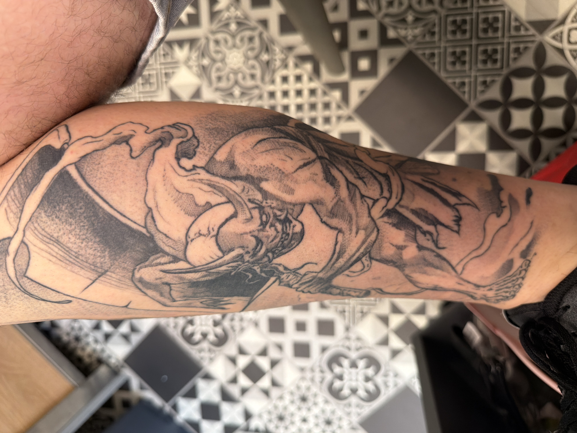 The image shows a person's lower leg with a large tattoo depicting a male figure wrestling a demonic creature. The tattoo is primarily in black ink and covers a significant portion of the lower leg. The wrestling figures are the main subject. The leg is positioned over a patterned tile floor. The tiles are black and white with geometric designs. The image is taken indoors. The environment includes a partial view of a table, and a shoe is visible, partially obscuring the edge of the leg. The person's skin tone is light, and they have noticeable leg hair above the tattoo. There is no discernible information about the location being in Sofia, Bulgaria.