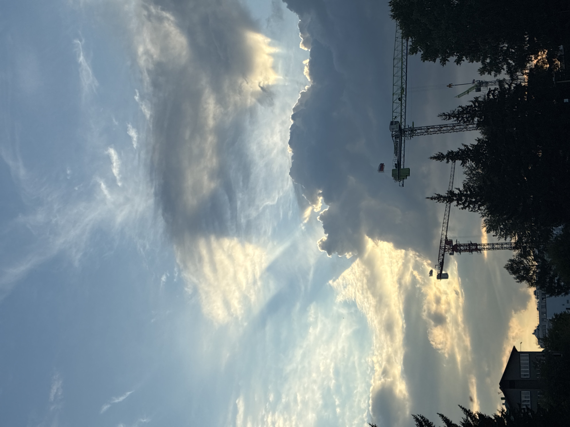The image depicts a construction site in Irkutsk, Russia, under a dramatic sky. The sky is filled with a mixture of dark, heavy clouds and lighter, wispy formations, with shafts of sunlight breaking through. Two large construction cranes are prominently featured, silhouetted against the sky, suggesting ongoing development. One crane, a tower crane, is a vibrant green and white, adorned with a Polish flag. Another, red and white tower crane, is partially visible on the left. In the foreground, the dark silhouettes of coniferous trees obscure the lower portion of the scene, with a glimpse of a building on the far left. The overall impression is one of industrial activity set against a natural, atmospheric backdrop. There are no people visible in the image.