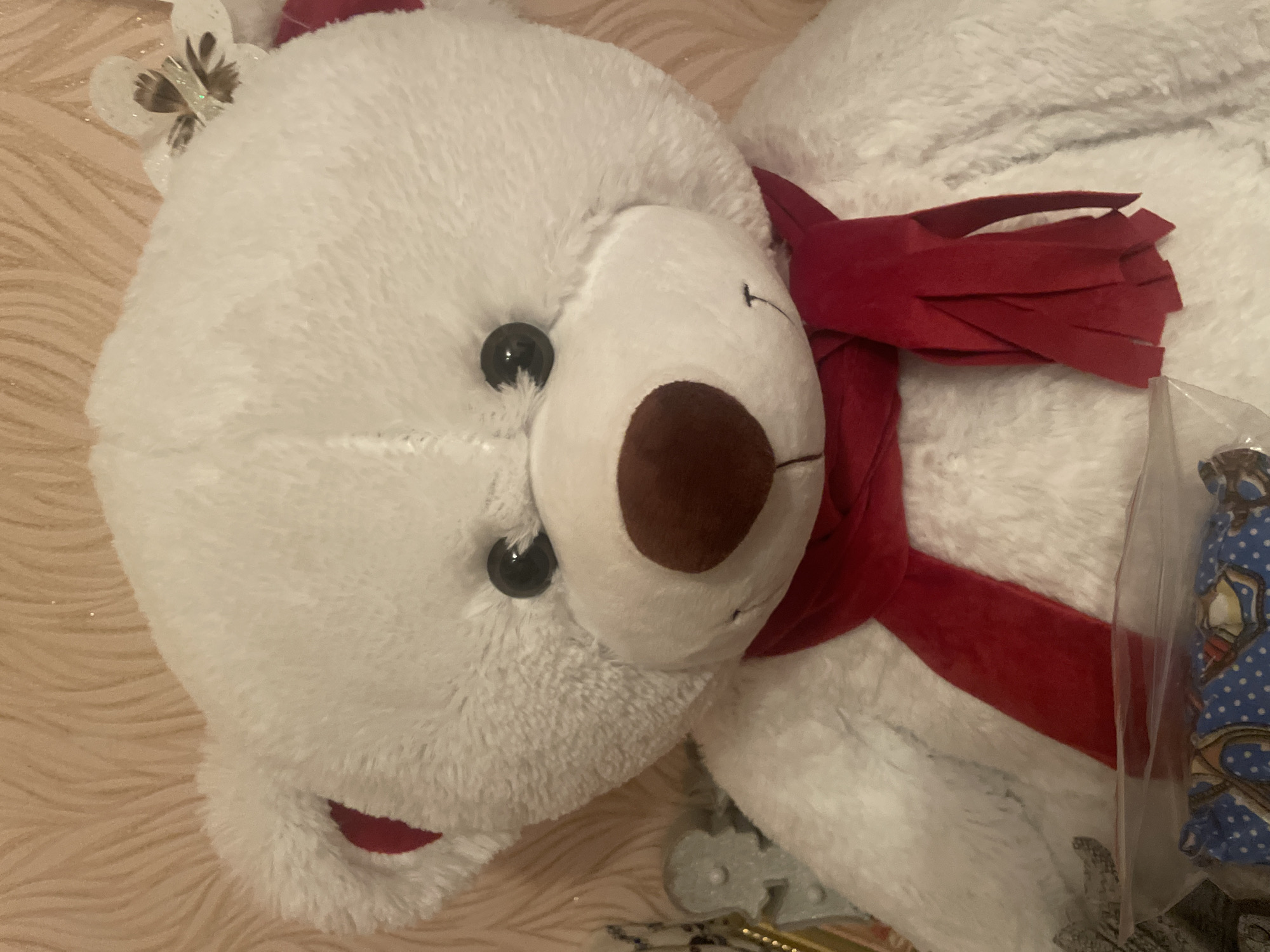 The media file shows a close-up, sideways view of a large, white, plush teddy bear. The bear is lying on a fuzzy, pale pink surface. The bear has black button eyes and a brown plush nose. It is wearing a bright red scarf, which is fringed at one end. A small, white, butterfly-shaped embellishment with glitter and darker details is visible on the bear's head, near its left ear. To the right of the bear, a clear plastic package is visible, containing a blue fabric with a white polka dot pattern and a repeating image of what appears to be a cowboy and horse. The lighting is warm, suggesting indoor illumination. No specific time of day, weather, or precise location can be determined from the image.