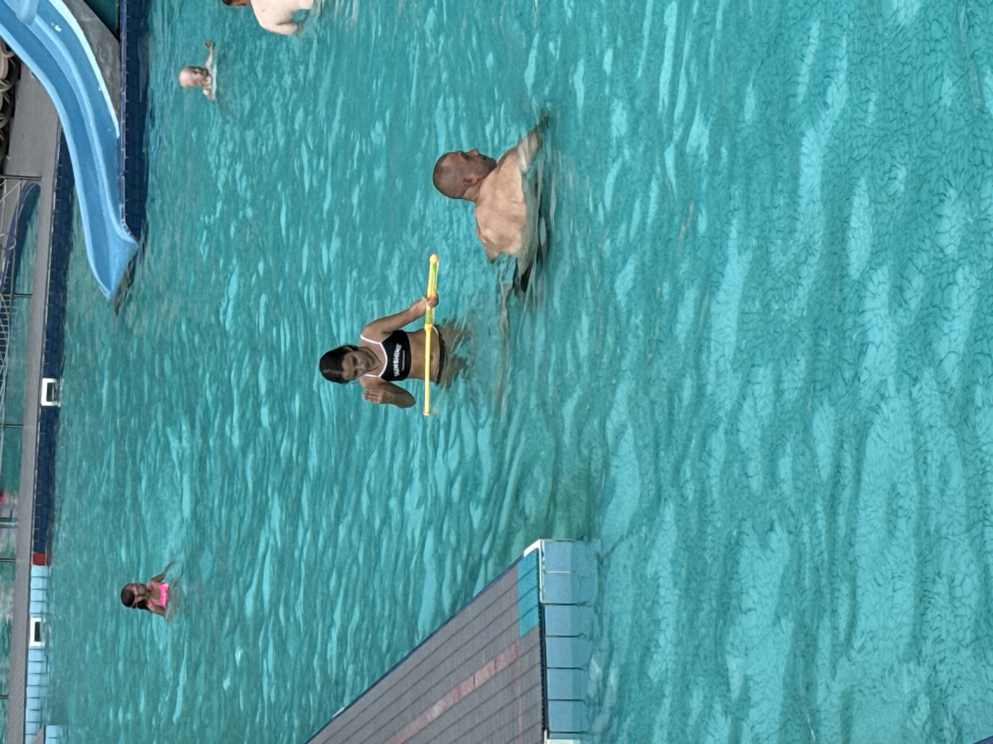This is an overhead, slightly tilted, medium shot of a swimming pool. There are at least four people visible in the pool. In the foreground, a young girl in a pink swimsuit and goggles is swimming. Further back, a woman in a black and white bikini is holding a yellow pool noodle. Behind her, a man with a shaved head is swimming. In the background, partially submerged, is another person. The water is a clear blue-green, and the surface is rippled. A blue water slide is visible on the left side of the frame, with a dark blue border. There are also some light blue and grey structures along the edge of the pool in the foreground. The lighting suggests it is daytime, and the weather appears to be clear. The scene is a recreational setting, likely an outdoor public pool. There is text visible on the woman's bikini, reading "SUNSHINE". The location is identified as Massandrivskyi village council, Ukraine.