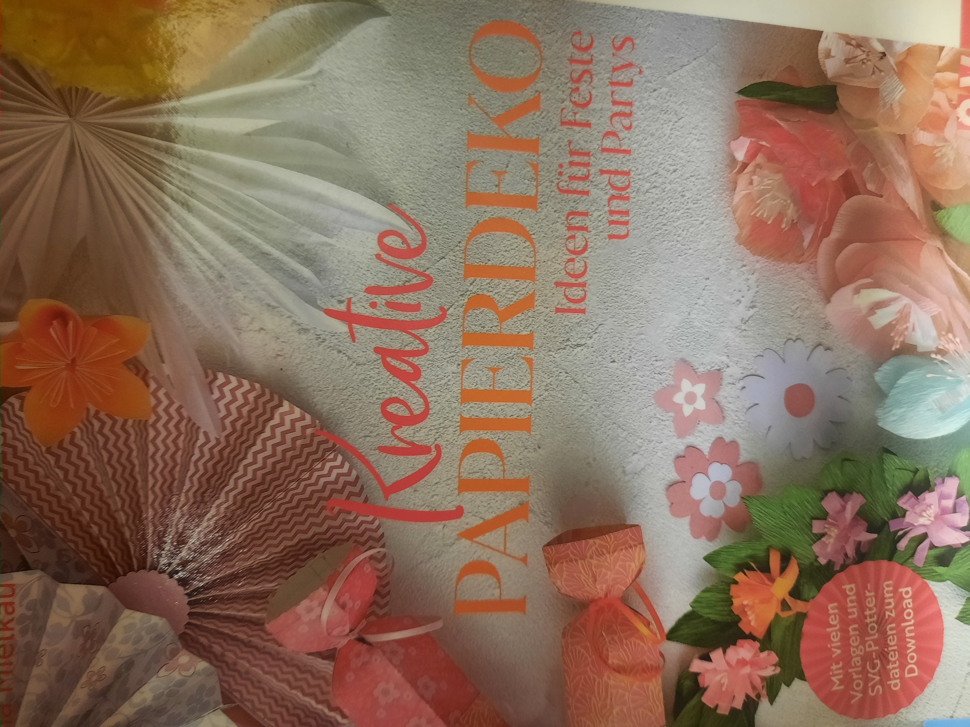 The image is a close-up, overhead shot of a craft magazine cover titled "Kreative PAPIERDEKO" (Creative PAPER DECOR). The background is a textured, light gray surface. The cover features various papercraft decorations, including folded paper fans, stylized paper flowers in shades of pink, orange, and light blue, and some gift-like paper structures with ribbon accents. The text "Ideen für Feste und Partys" (Ideas for celebrations and parties) is visible below the main title. In the bottom left corner, a circular red stamp reads "Mit vielen Vorlagen und SVG-Plotter-dateien zum Download" (With many templates and SVG plotter files for download). The magazine appears to be a lifestyle or hobby publication. There is no indication of the city of Rosenheim, Germany in the image.