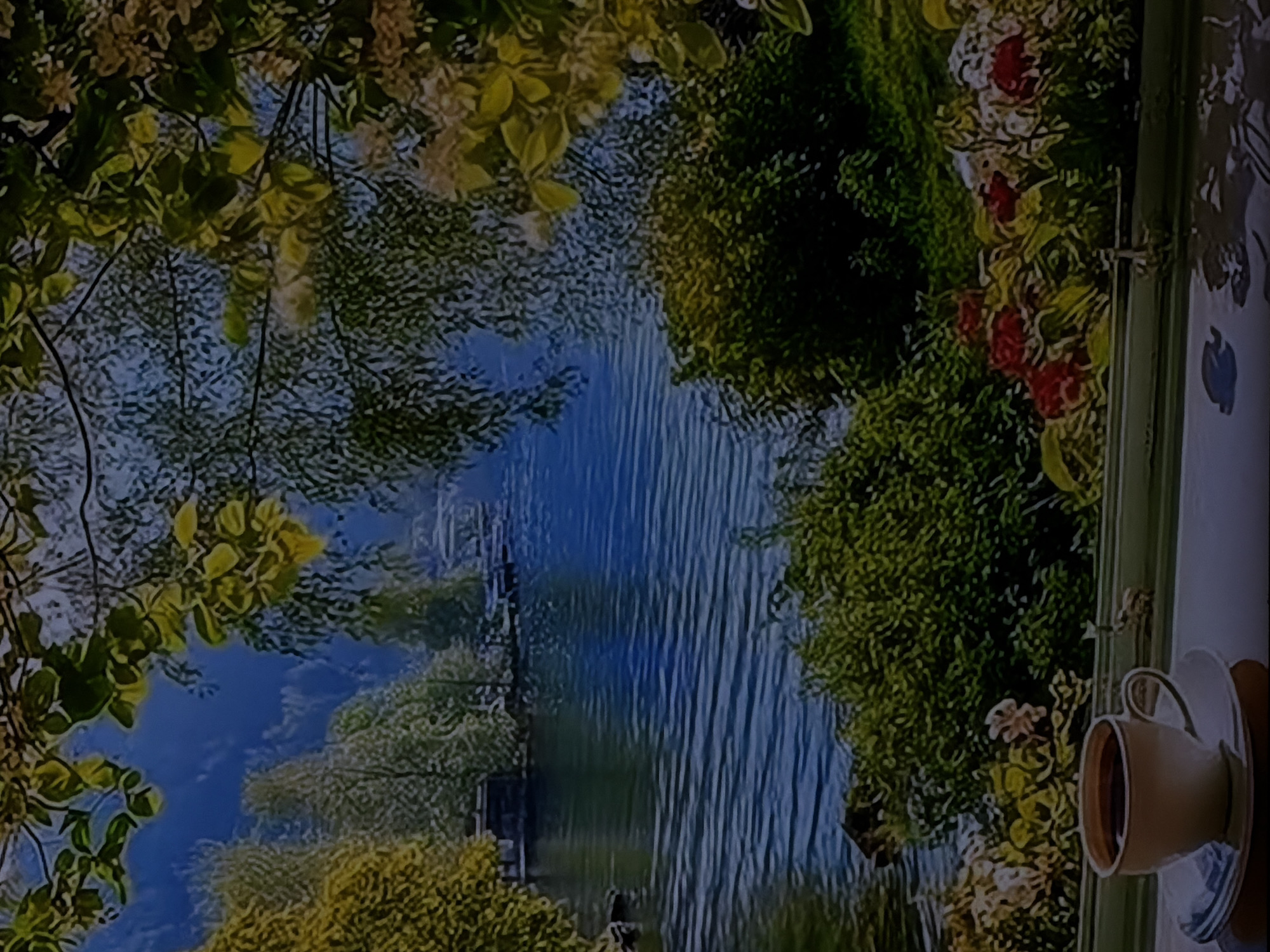 The image displays a serene landscape scene, likely viewed from an indoor setting. At the forefront, on the right side of the image, a white ceramic cup with a golden rim rests on a matching saucer, filled with a dark liquid, presumably coffee or tea. This arrangement suggests an individual is enjoying a beverage while observing the view.
The main scene features a tranquil body of water, possibly a river or lake, running through the center. Its surface shows subtle ripples or reflections, creating vertical textures. The water is bordered by lush, abundant vegetation on both banks. The foliage varies in shade from dark, dense green bushes to lighter, yellow-green leafy trees. Overhanging branches with delicate leaves frame the upper part of the vista. On the right bank, interspersed among the green foliage, are patches of flowering plants with vibrant red and white blossoms. A dark structure, possibly a small dock or pier, is faintly visible on the left bank of the water.
The overall setting combines an inviting indoor space, hinted at by the cup and saucer on what appears to be a windowsill or table, with an idyllic outdoor natural environment. The scene is illuminated by soft, natural daylight, suggesting a pleasant morning or afternoon. The aesthetic quality of the natural landscape, with its rich colors and somewhat stylized textures, could indicate that it is a mural, a high-quality print, or an actual view from a window. There are no specific visual cues within the image itself to definitively identify the location as Sofiivska Borshchahivka, Ukraine; it presents a generic yet picturesque natural vista. No text is visible in the image.