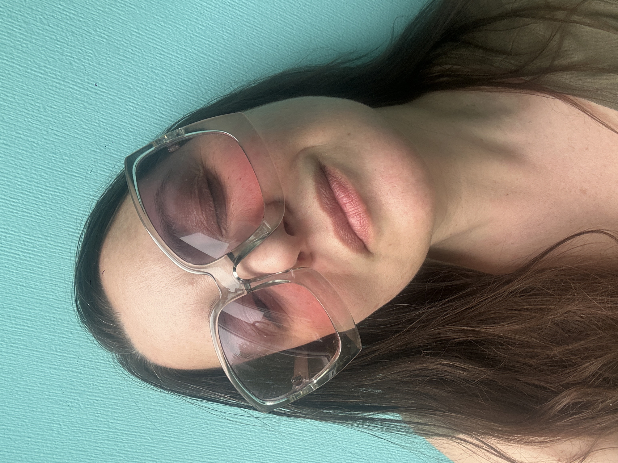 A woman with long brown hair is lying on a light blue surface wearing sunglasses. The image does not contain enough information to determine the location as Riga, Latvia.