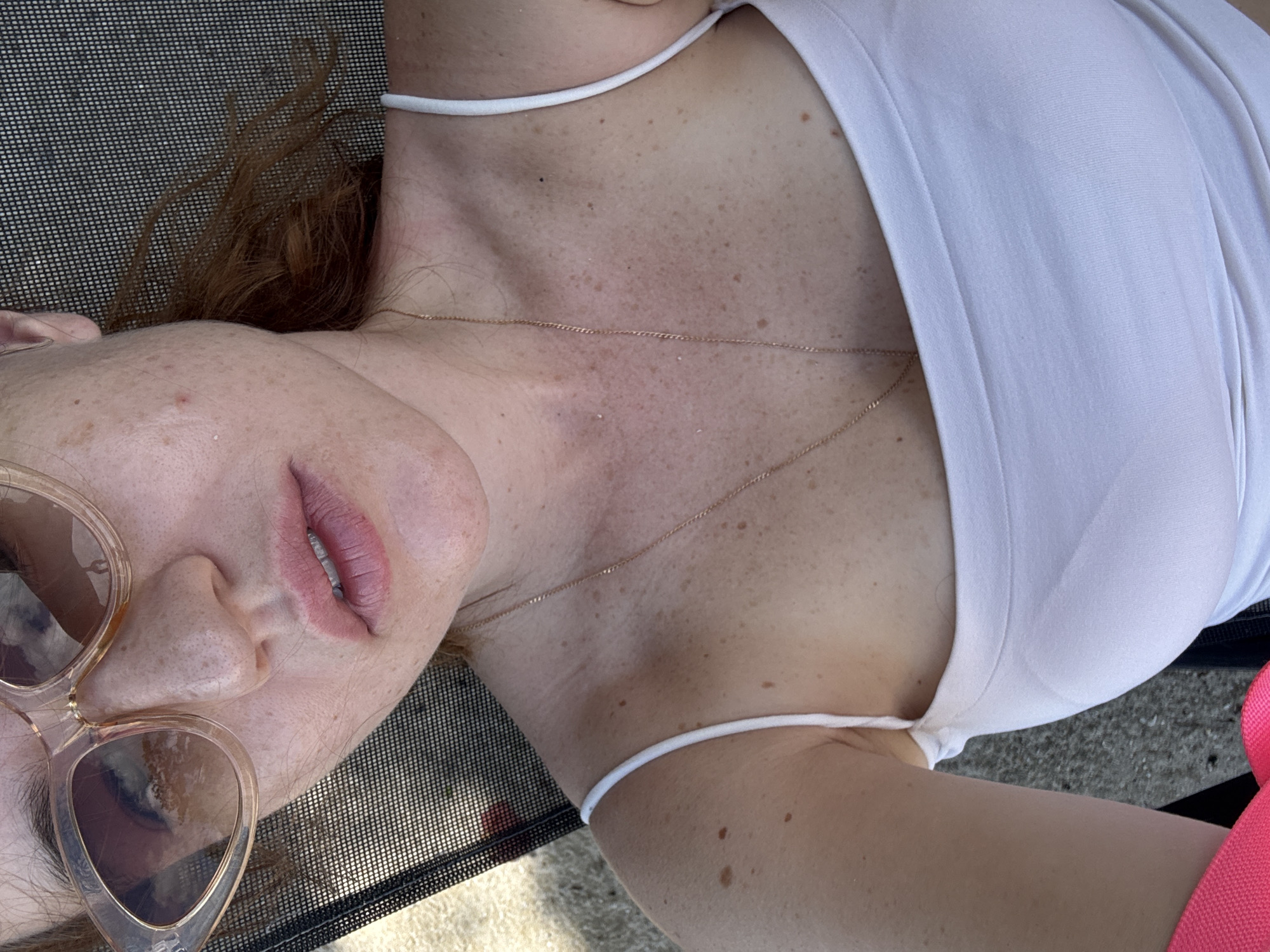 A close-up, overhead shot shows a person lying on their back on a textured dark surface, possibly a lounge chair. Their head is angled downwards, with their face partially obscured by large, light-colored sunglasses. Their lips are slightly parted, revealing white teeth. The person has fair skin with numerous freckles across their face and chest. A delicate gold chain necklace rests on their collarbone and chest. They are wearing a white tank top with thin straps. The background consists of the dark textured surface and a lighter, possibly concrete or sand, ground. The lighting suggests it is daytime, and the overall mood appears relaxed, possibly indicating a sunny day spent outdoors. The scene is set in Baan Plai Laem, Thailand.