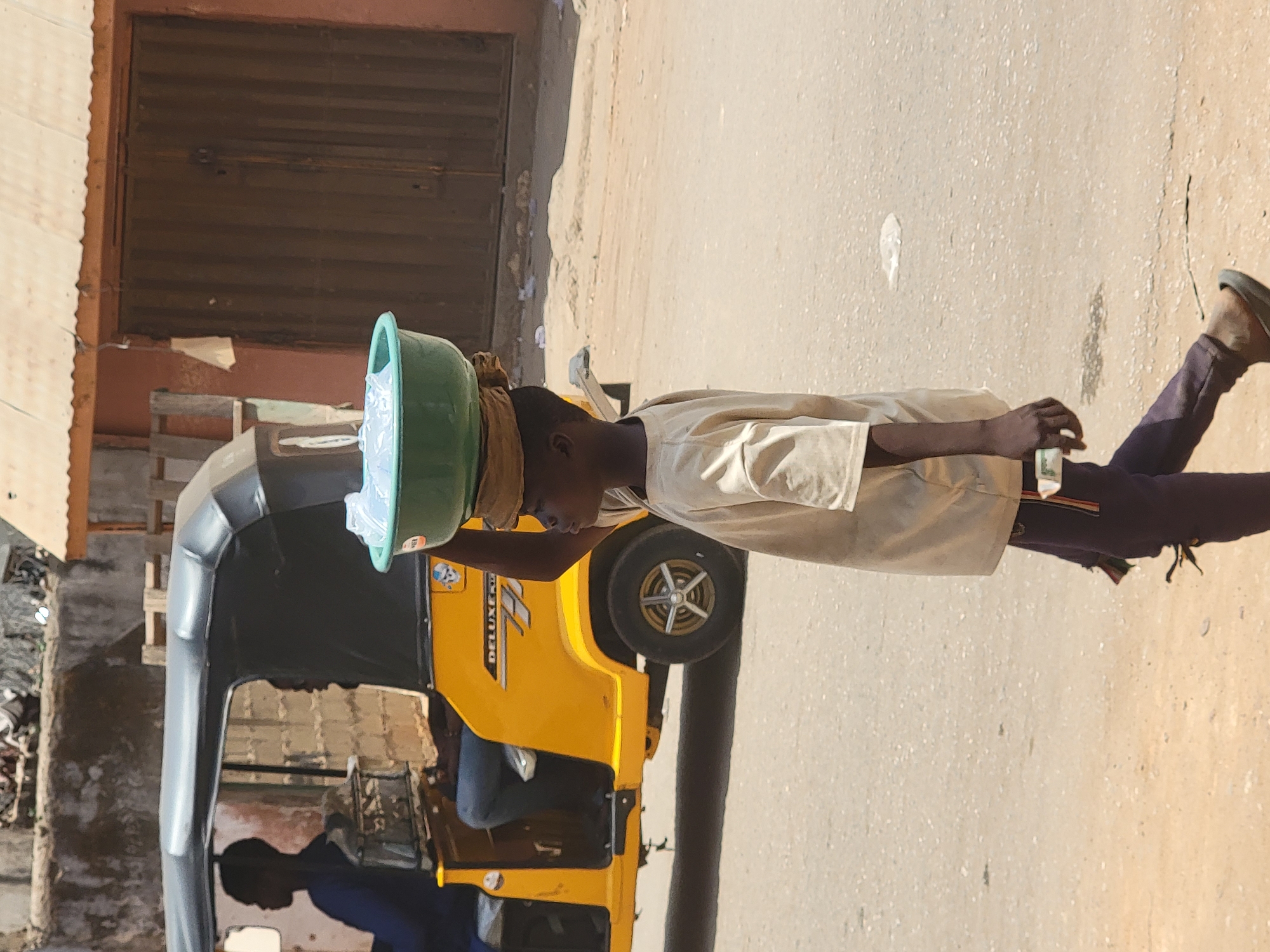 In Maiburiji, Nigeria, an individual wearing a beige shirt and dark pants carries a green basin filled with transparent bags of water on their head. They are walking along a paved road beside a yellow and black three-wheeled vehicle. The vehicle is parked near a building with a closed brown metal door. The environment appears to be a street scene during daylight.