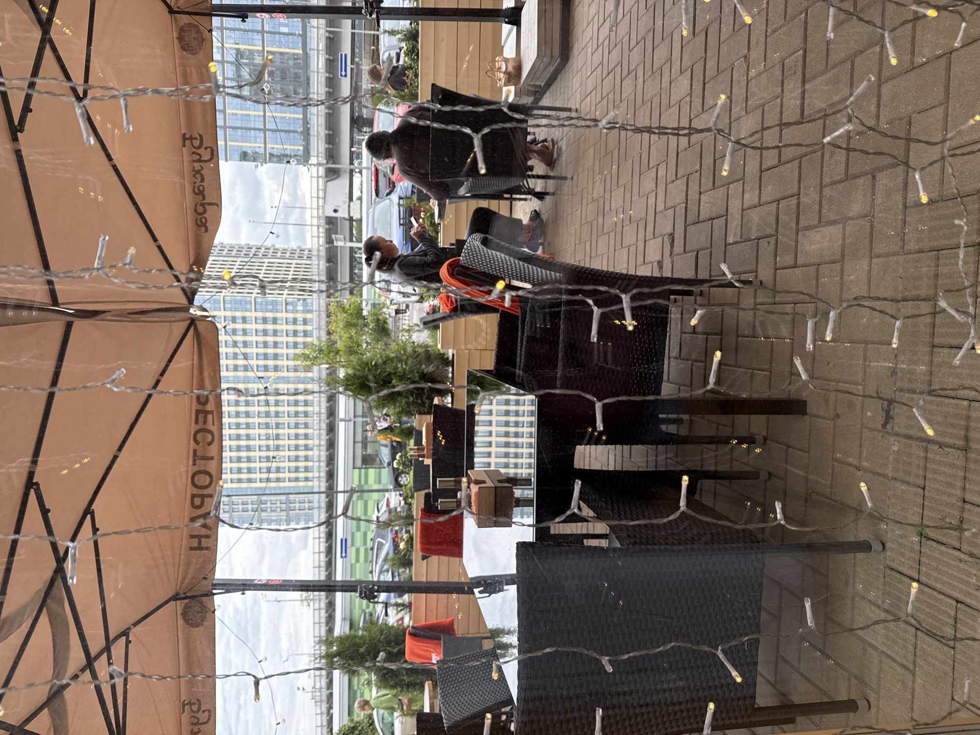 The image depicts an outdoor cafe or restaurant patio, viewed through a glass window with decorative string lights prominently strung across the foreground. The scene is composed of several dark wicker-style chairs and tables with highly reflective surfaces, arranged on a paved ground. Large, beige umbrellas provide shade over the seating areas.
Several people are seated at the tables. In the mid-ground, a person with short hair is engrossed in their smartphone. Further back, another individual is seated with a bag placed on an adjacent surface. Other patrons are partially visible in the distance. Potted plants and shrubs are used as decorative elements or dividers within the patio.
Beyond the immediate seating area, a street or parking lot is visible with cars. The background features modern, multi-story buildings with numerous windows, indicating an urban setting. The lighting suggests it is daytime, possibly under an overcast sky, as evidenced by the soft illumination and reflections.
A notable detail is the Cyrillic text "Ресторан" (Restoran), meaning "Restaurant," clearly visible on one of the large beige umbrellas. Reflections of the surrounding environment, including buildings and the sky, are noticeable on the table surfaces and the glass through which the photograph was captured. The string lights, with their small, clear bulbs, contribute to the cafe's ambiance. The location is Moscow, Russia.